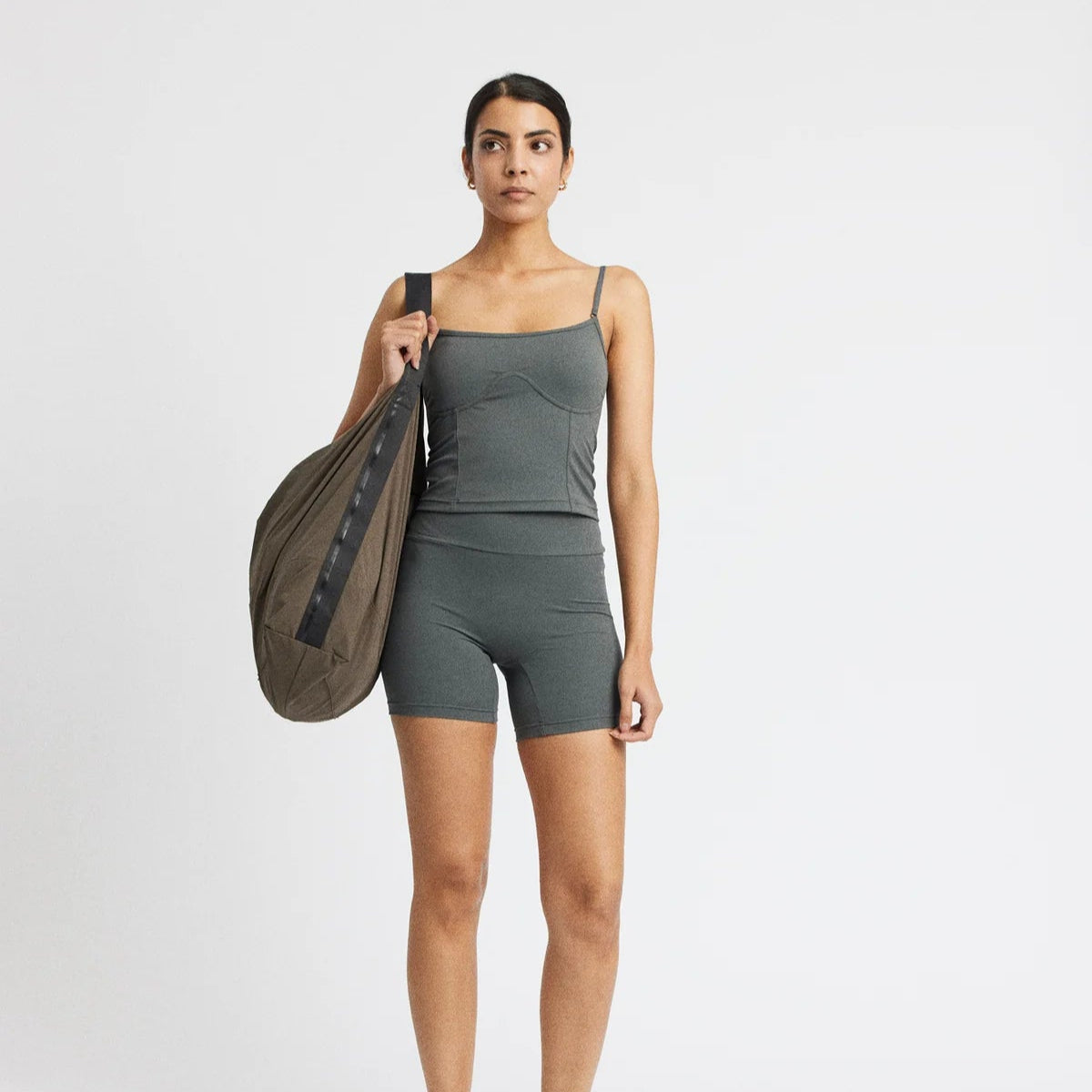 Frau in grauem Sportoutfit hält eine große braune Tasche, steht vor einem neutralen Hintergrund.