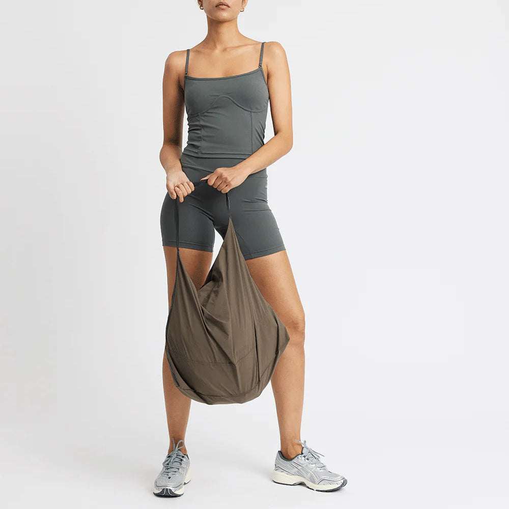Frau in grauem Sportoutfit hält eine braune Tasche, stehend vor weißem Hintergrund.