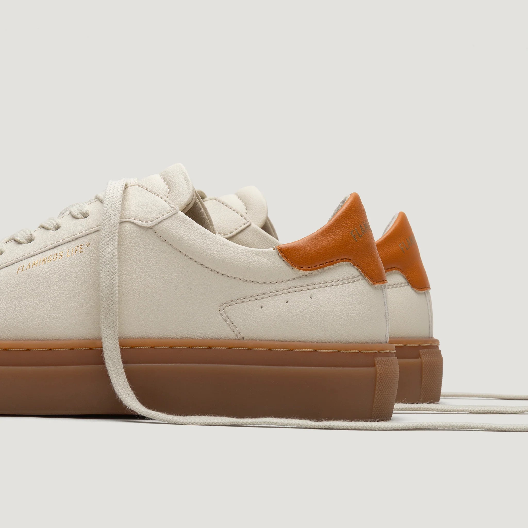 Weiße Sneaker mit brauner Sohle und orangen Akzenten, seitlich und teilweise von hinten abgebildet.