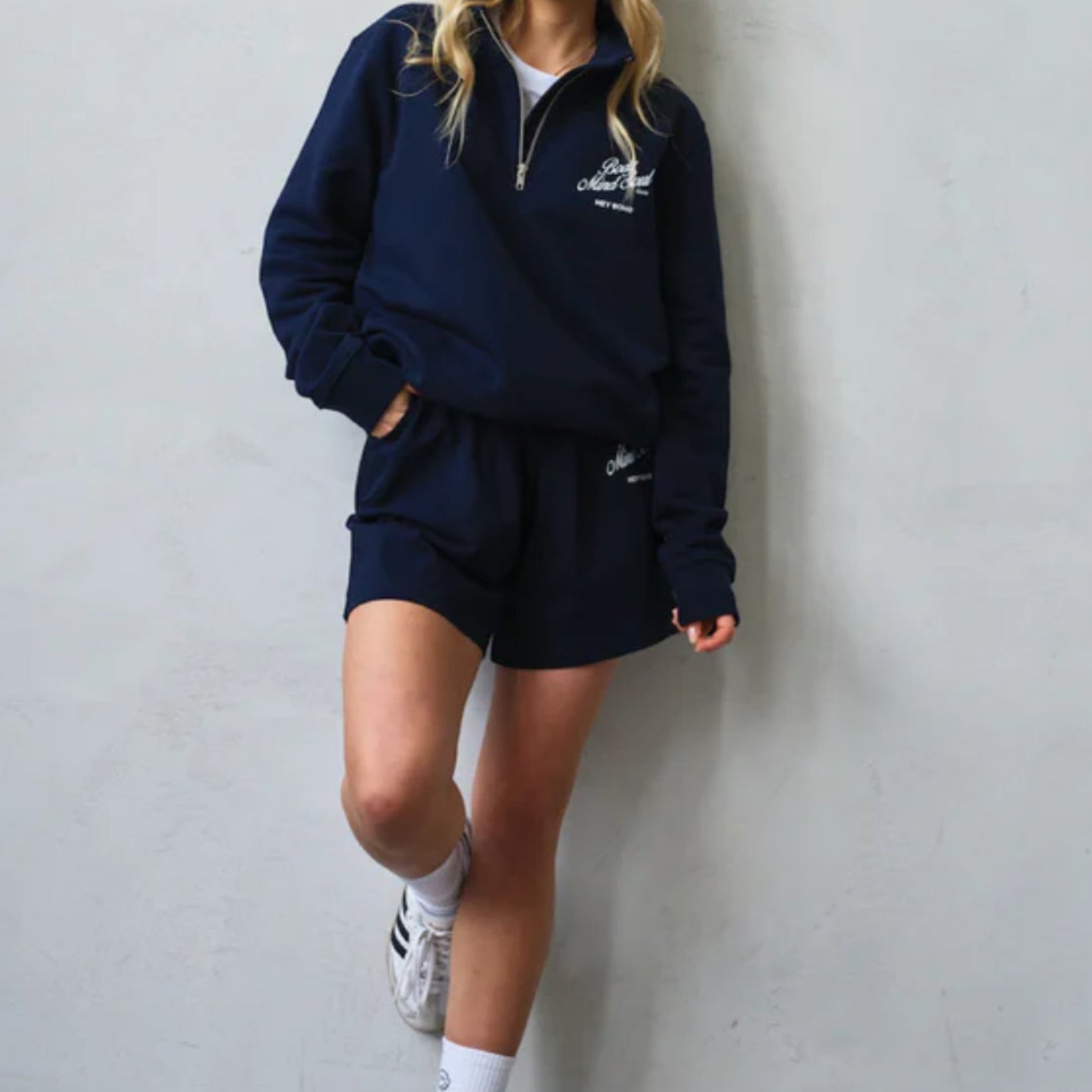 Person lehnt an einer Wand und trägt einen dunkelblauen Trainingsanzug mit Shorts und weißen Adidas-Sneakern.