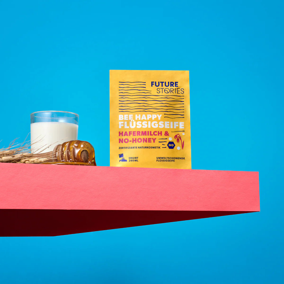 Eine Packung Future Stories Bee Happy Flüssigseife steht auf einem rosa Regal neben einem Glas Milch, Weizen und Honigkeksen, alles vor einem hellblauen Hintergrund.