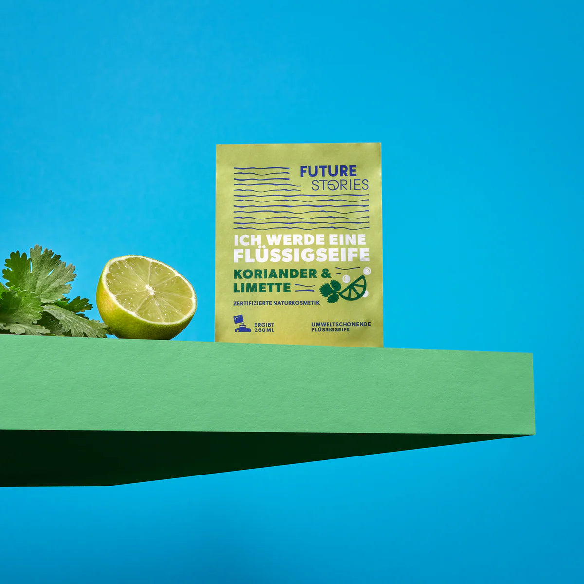 Eine Packung Future Stories Flüssigseife mit der Aufschrift Koriander & Limette steht aufrecht in einem grünen Regal, neben einer halben Limette und Korianderblättern, vor einem hellblauen Hintergrund.