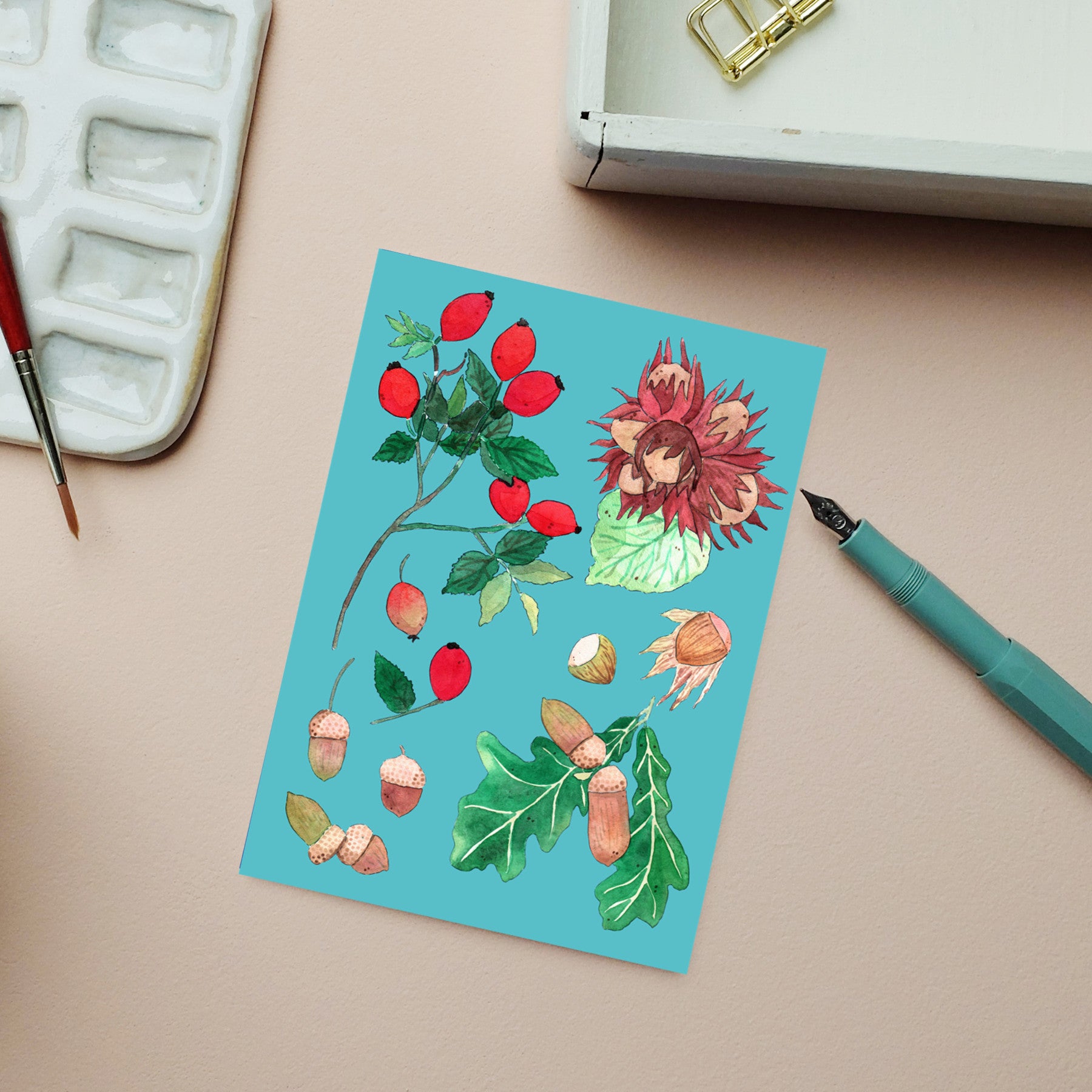 Auf einer Karte auf dem Schreibtisch sind Aquarellbilder von Hagebutten, Eicheln, einer geöffneten Kastanie, Blättern und einem Füllfederhalter zu sehen, daneben ein Pinsel, eine Farbpalette und ein weißer Kasten.