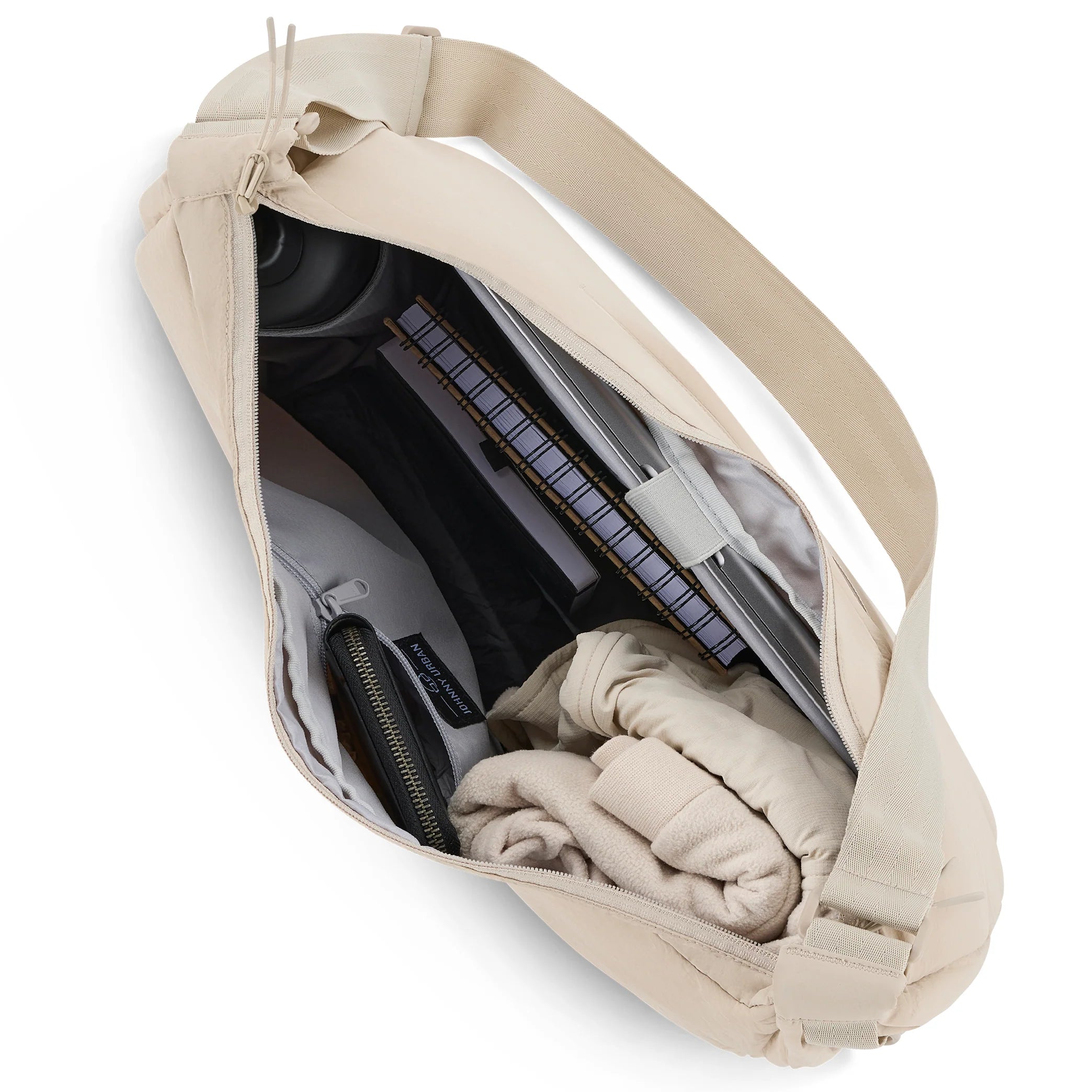 Eine beigefarbene Umhängetasche mit geöffnetem Reißverschluss, in der sich ein Spiralnotizbuch, eine Wasserflasche, eine Tasche mit Reißverschluss und gefaltete helle Kleidung befinden.