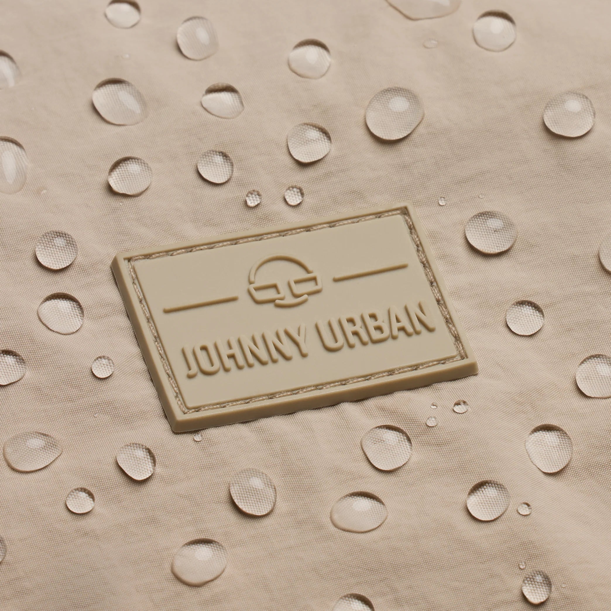 Ein beigefarbenes Stoffetikett mit JOHNNY URBAN und einem Logo ist mit Wassertropfen bedeckt, was die wasserabweisende Qualität des Materials unterstreicht.