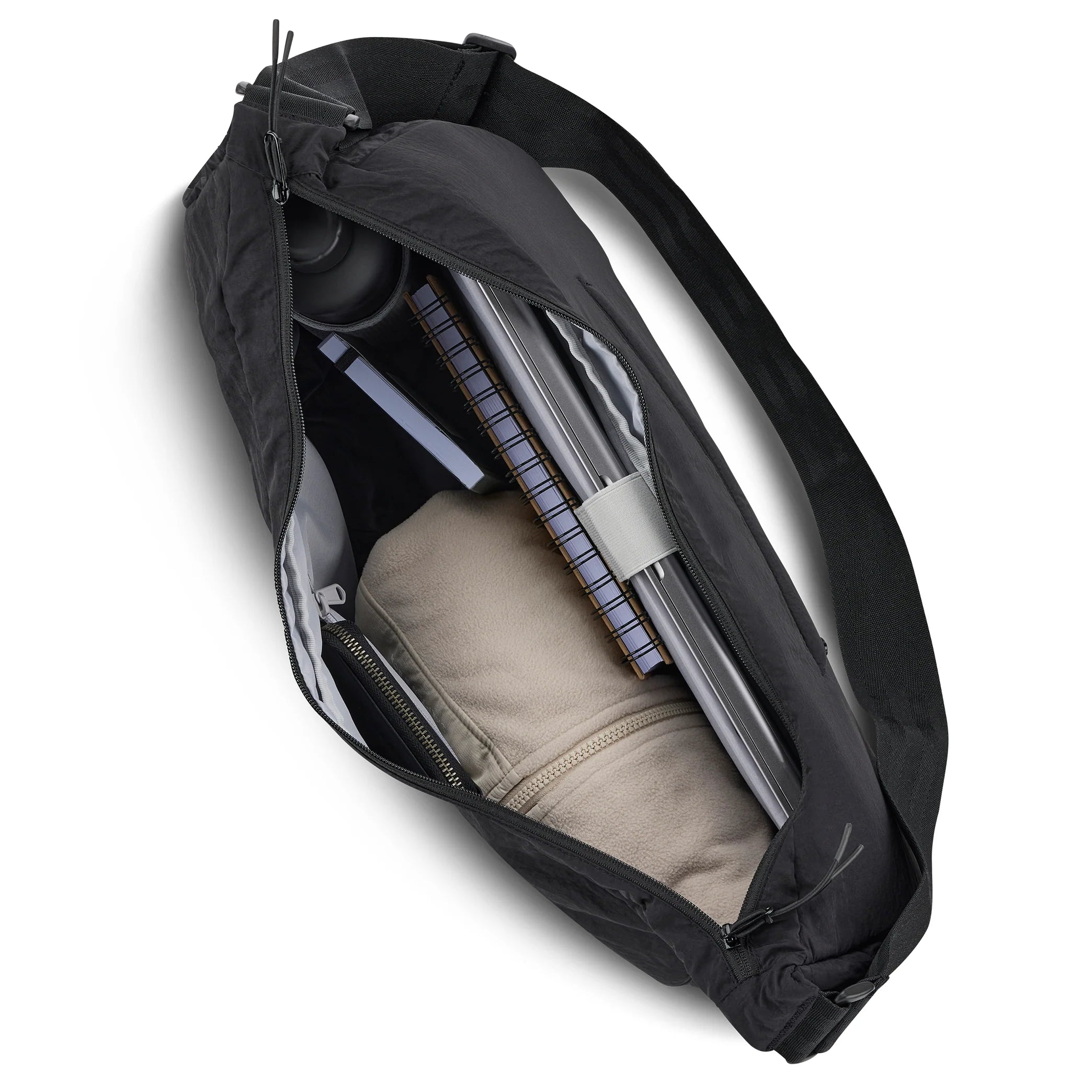 Eine schwarze Umhängetasche, die von oben betrachtet wird und deren Reißverschluss geöffnet wurde, um einen beigen Kapuzenpullover, ein Spiralnotizbuch, einen Laptop, eine Wasserflasche und andere kleine Gegenstände zum Vorschein zu bringen, die in ihrem Inneren fein säuberlich angeordnet sind.