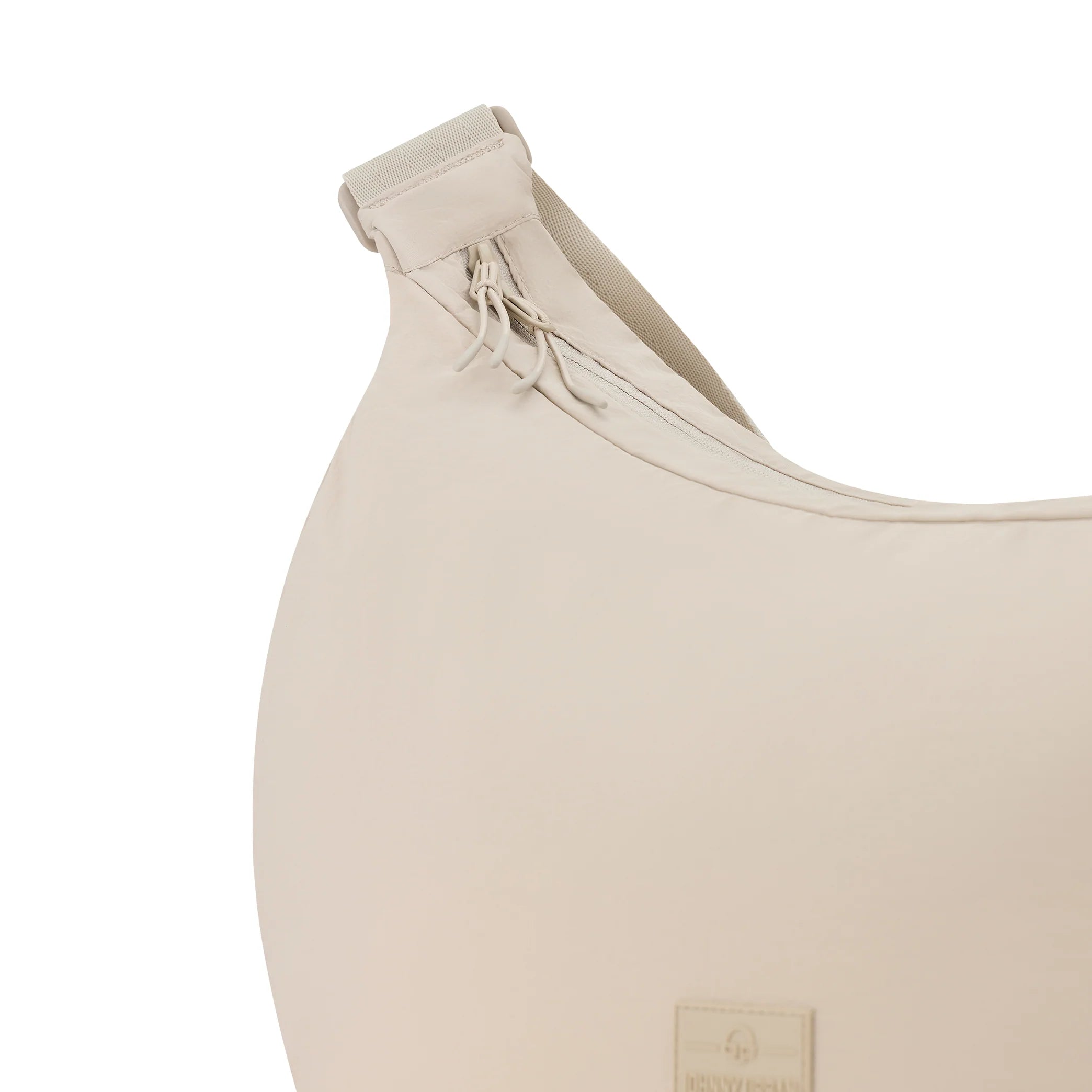 Nahaufnahme einer hellbeigen Stofftasche mit geschwungener Form, die einen Reißverschluss mit zwei Zuglaschen im oberen Bereich und eine Stoffschlaufe an der Ecke aufweist. Ein kleiner rechteckiger Logoaufnäher ist teilweise am Boden sichtbar.