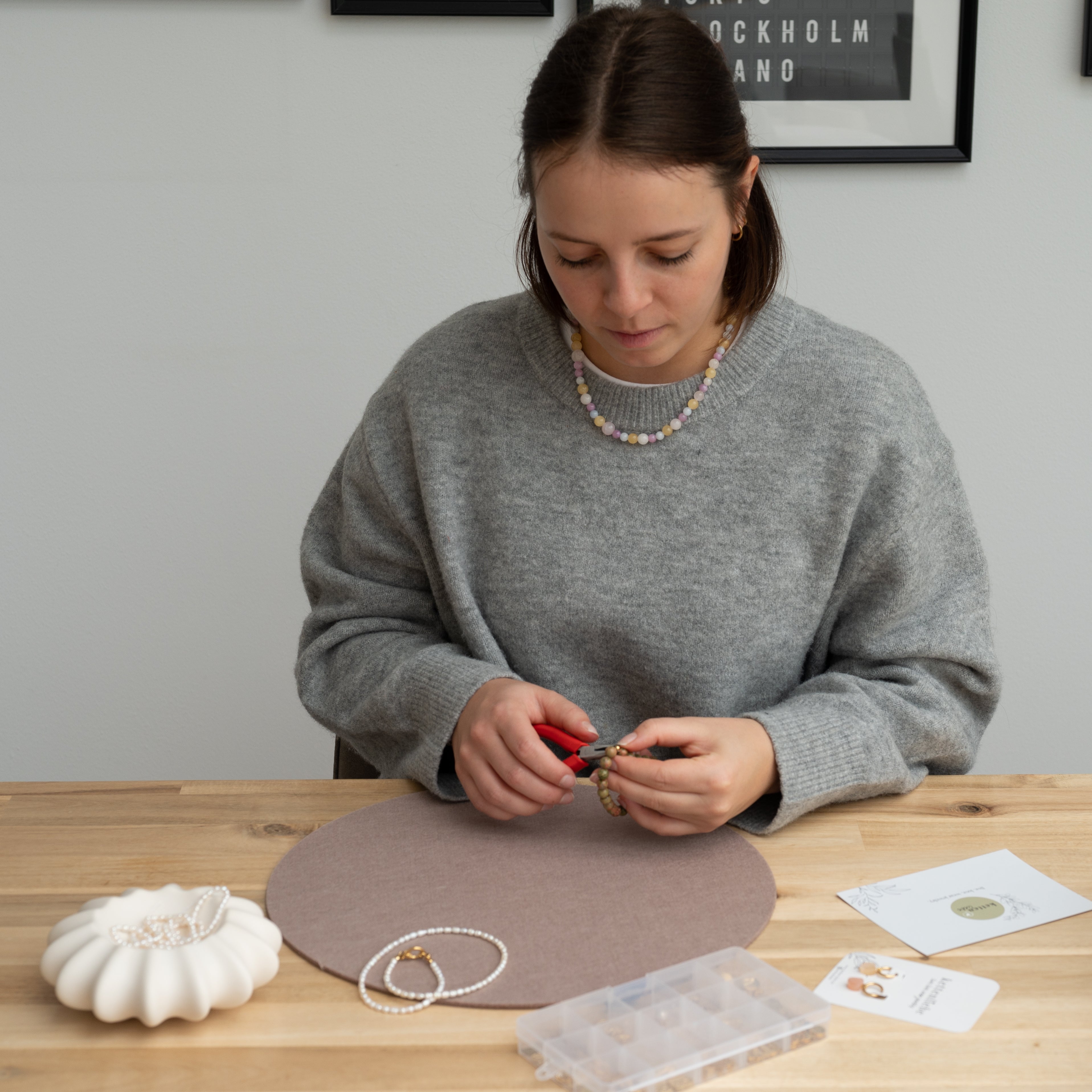 Eine Frau in einem grauen Pullover sitzt an einem Tisch und bastelt mit einer Zange Schmuck. Auf dem Tisch vor ihr liegen Perlen, eine Perlenkette und eine Organizerbox.