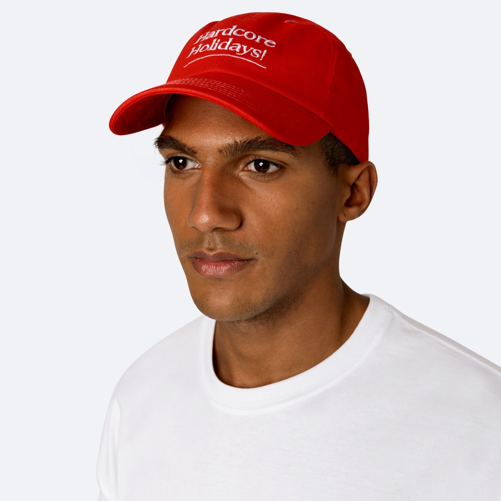 Eine Person, die eine leuchtend rote Baseballmütze mit der Aufschrift Hardcore Holidays! in weißer Schrift und ein schlichtes weißes T-Shirt trägt und vor einem neutralen Hintergrund leicht zur Seite schaut.