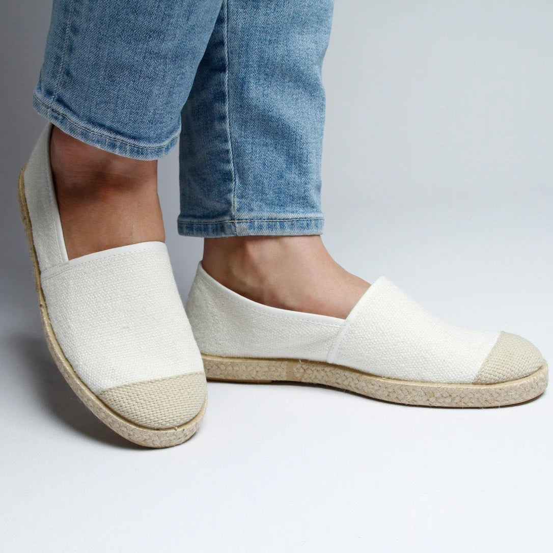 Person trägt weiße Slip-on-Schuhe und hellblaue Jeans auf einem hellen Hintergrund.