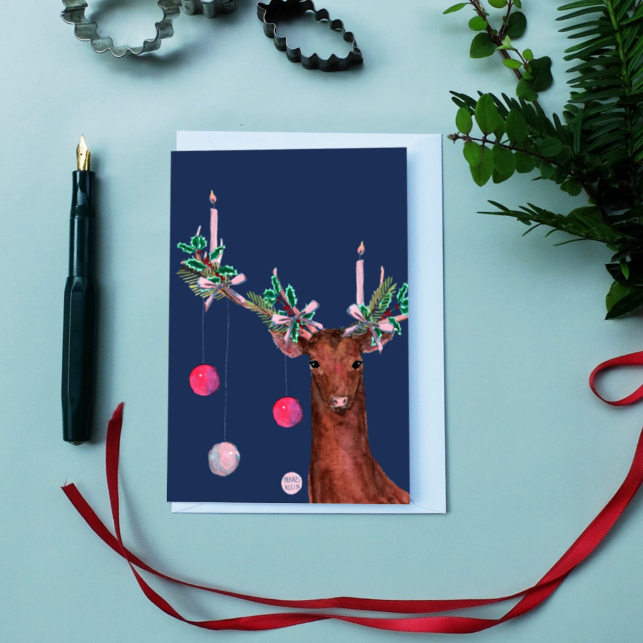 Weihnachtskarte mit Rentier, dessen Geweih mit Kerzen, Girlanden und Kugeln dekoriert ist, umgeben von Pflanzen und Stift.