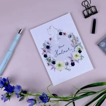 Eine Grußkarte mit einem Blumenkranz-Design liegt auf einem rosa Hintergrund neben einem Füller und Blumen.