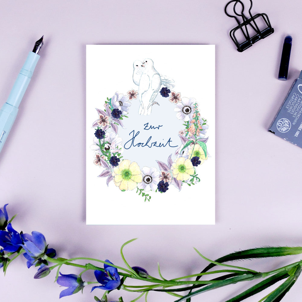 Hochzeitskarte mit Blumenkranz und Tauben, umgeben von Schreibutensilien auf einem hellvioletten Hintergrund.