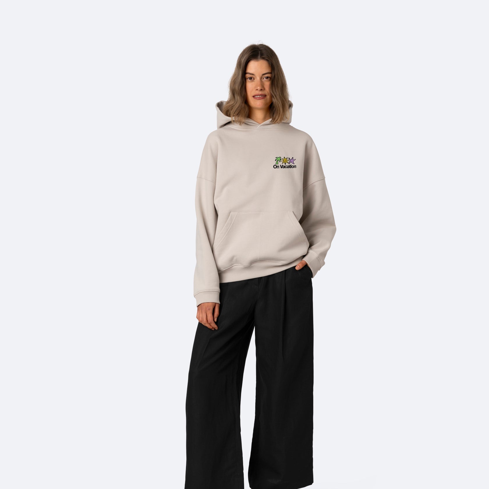 Eine Person steht vor einem schlichten, hellen Hintergrund und trägt einen beigen Kapuzenpullover mit einer kleinen bunten Grafik auf der Brust, eine weite schwarze Hose und schwarze Sandalen.