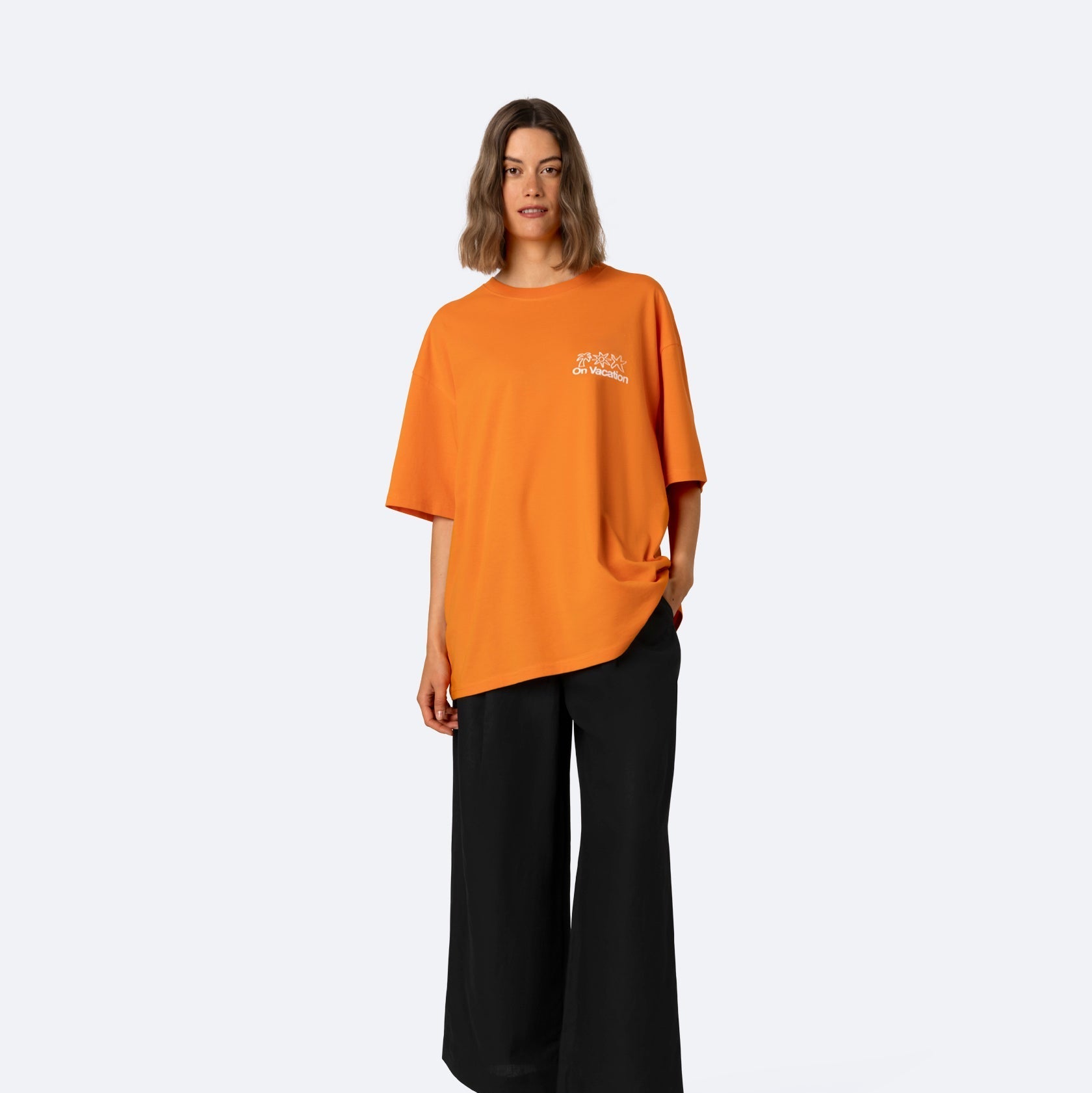 Eine Frau steht vor einem schlichten, hellen Hintergrund. Sie trägt ein übergroßes orangefarbenes T-Shirt mit weißer Aufschrift auf der Brust, eine weite schwarze Hose und schwarze Sandalen. Ihr Haar ist schulterlang und sie hat eine Hand in der Tasche.