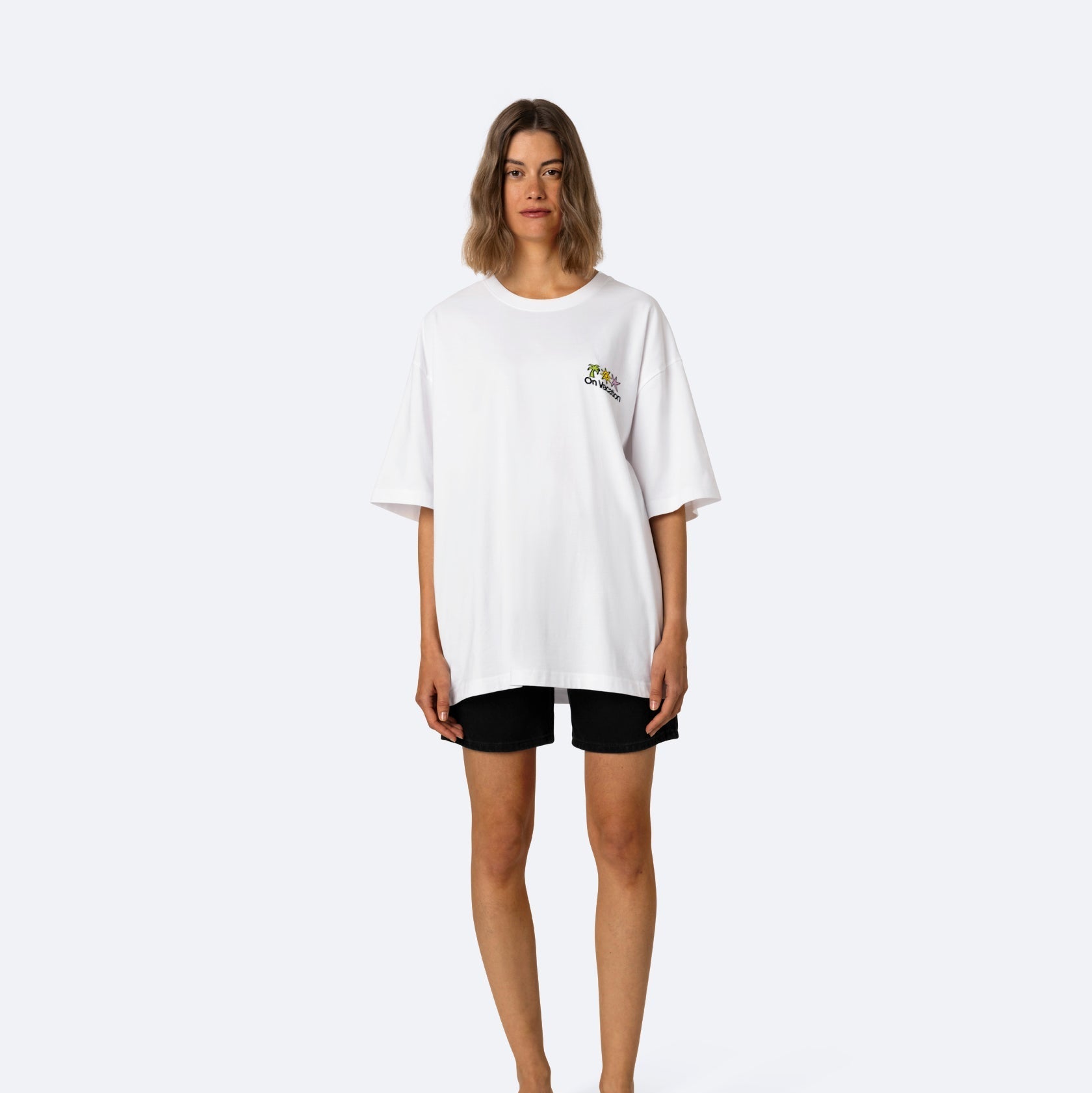 Eine Frau mit schulterlangen Haaren trägt ein übergroßes weißes T-Shirt mit einer kleinen Grafik auf der Brust, schwarze Shorts und schwarze Plateausandalen und steht vor einem schlichten hellgrauen Hintergrund.