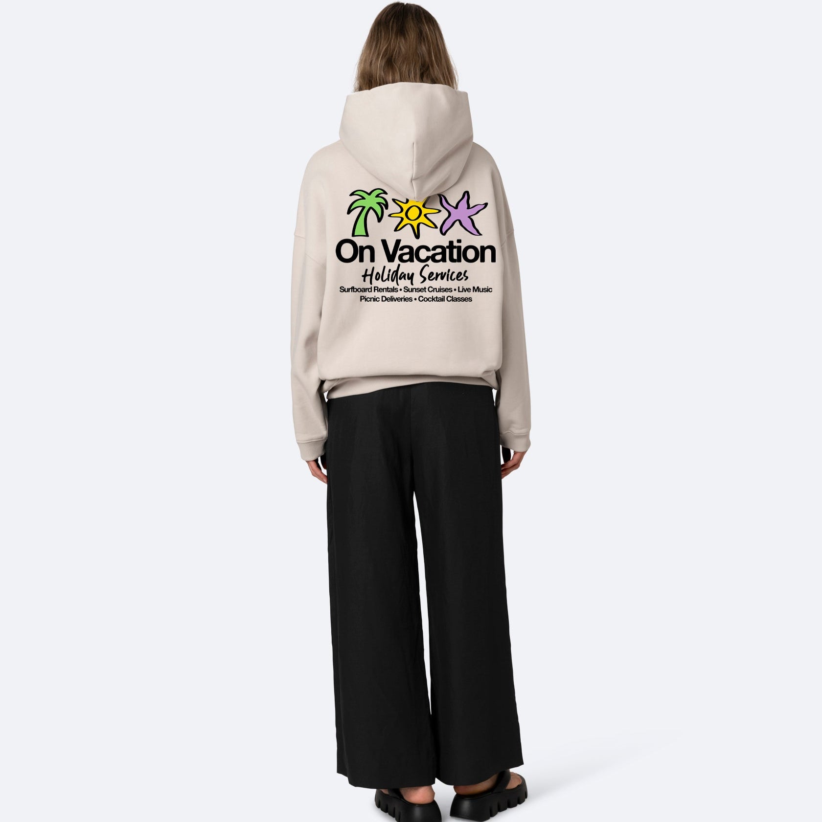 Eine Person steht mit dem Gesicht zur Wand und trägt einen beigen Kapuzenpullover mit bunten Palmen und Seesternen und der Aufschrift On Vacation Holiday Services auf dem Rücken, dazu eine weite schwarze Hose und schwarze Sandalen.
