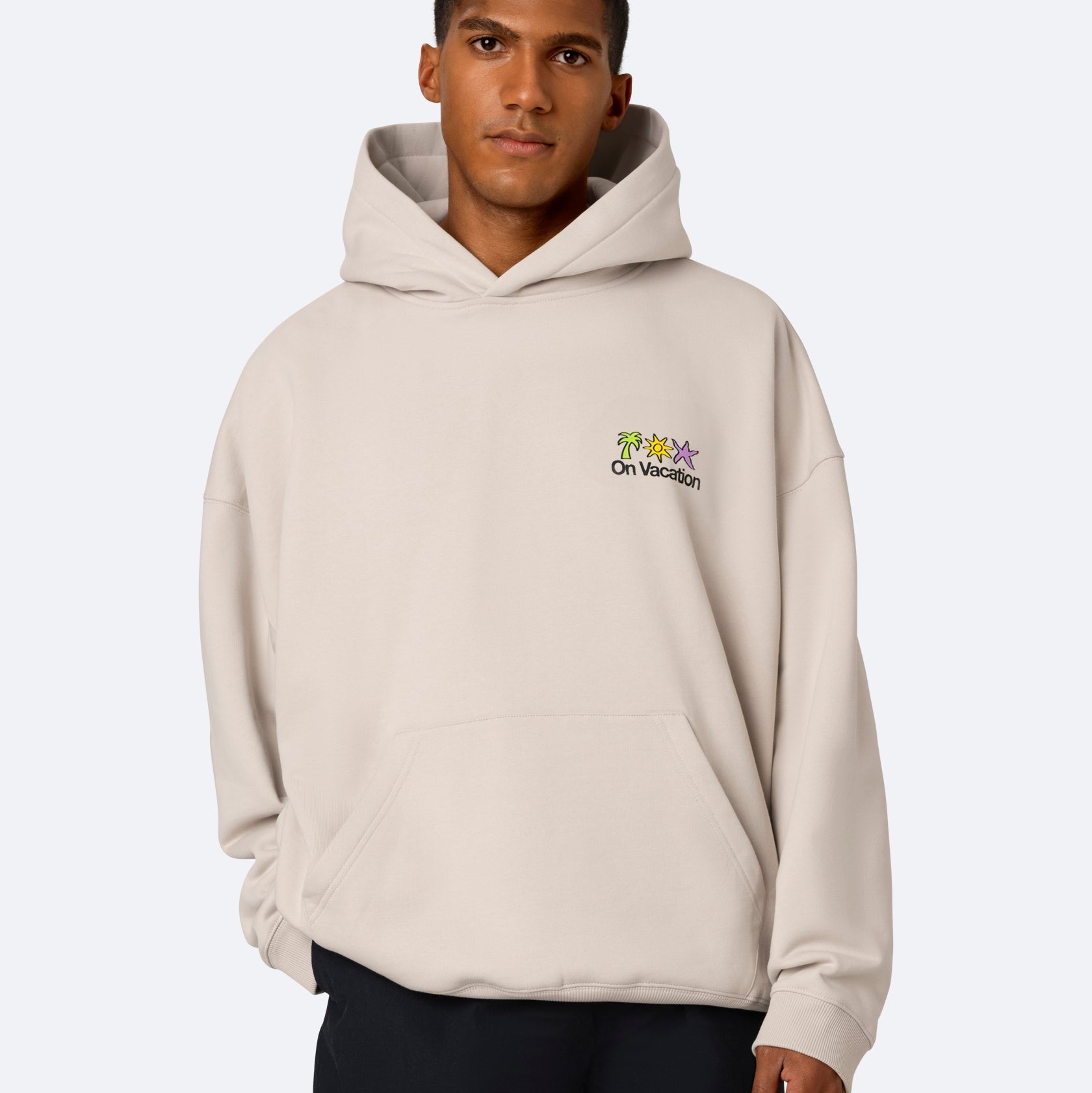 Ein junger Mann trägt ein beiges Kapuzensweatshirt mit bunten Cartoon-Figuren und der Aufschrift On Vacation auf der Brust vor einem schlichten hellen Hintergrund.