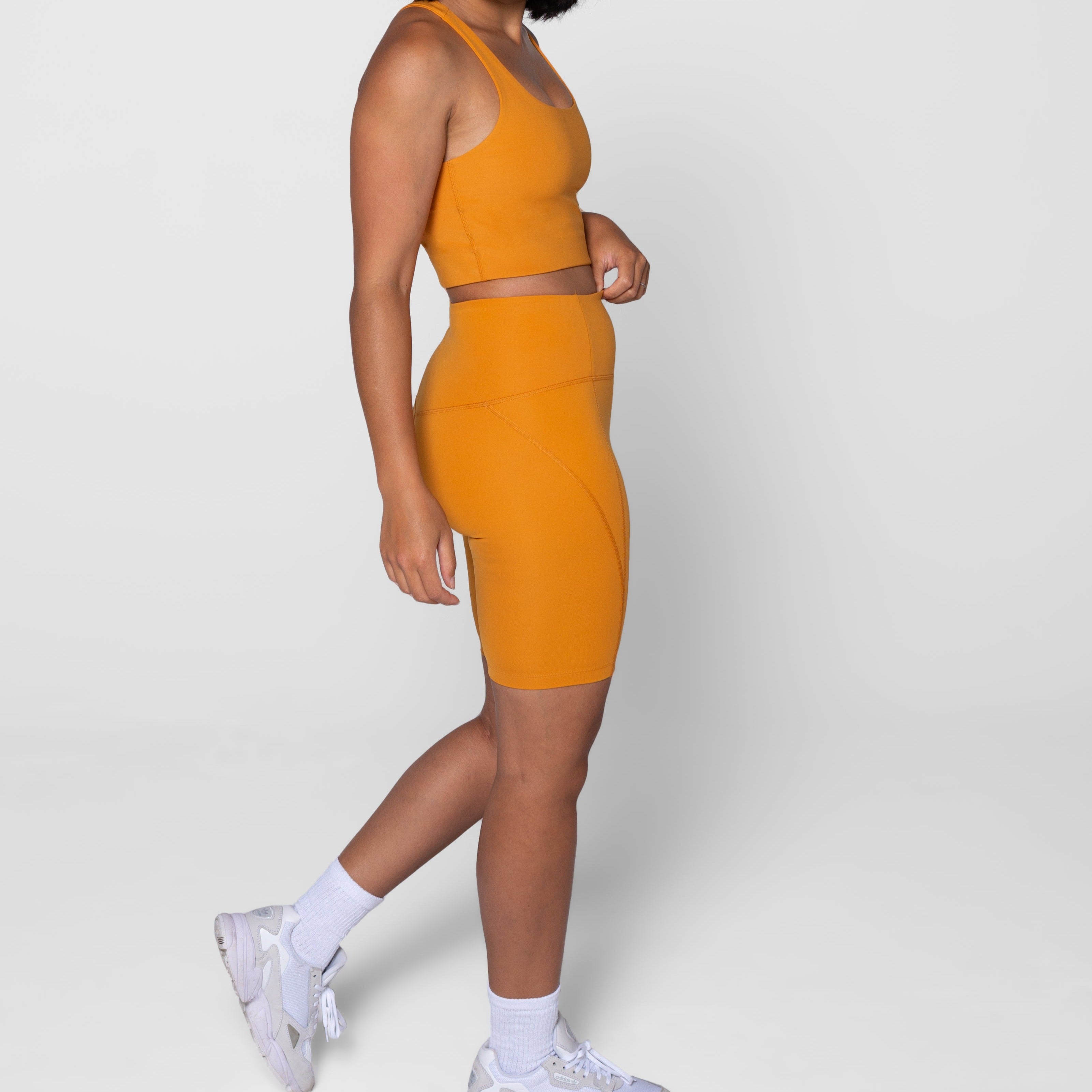 Person in orange athletic outfit with matching top and cycling shorts, wearing white sneakers on a gray background.