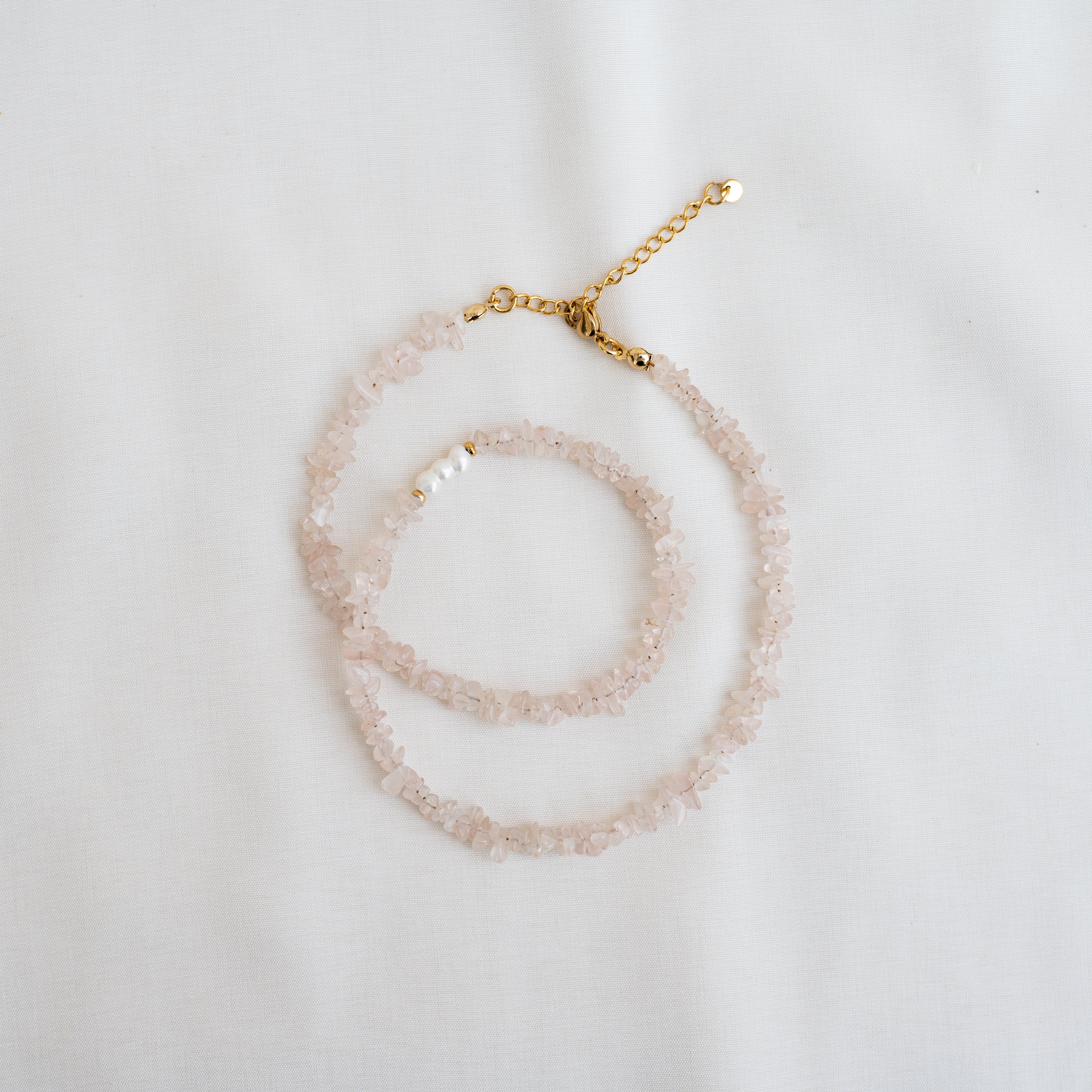 Eine zarte Halskette aus hellrosa Edelsteinchips mit einer goldenen Kette und einem goldenen Verschluss, die auf einem weißen Stoffhintergrund aufgewickelt sind. In der Nähe des Verschlusses befinden sich einige weiße Perlen.