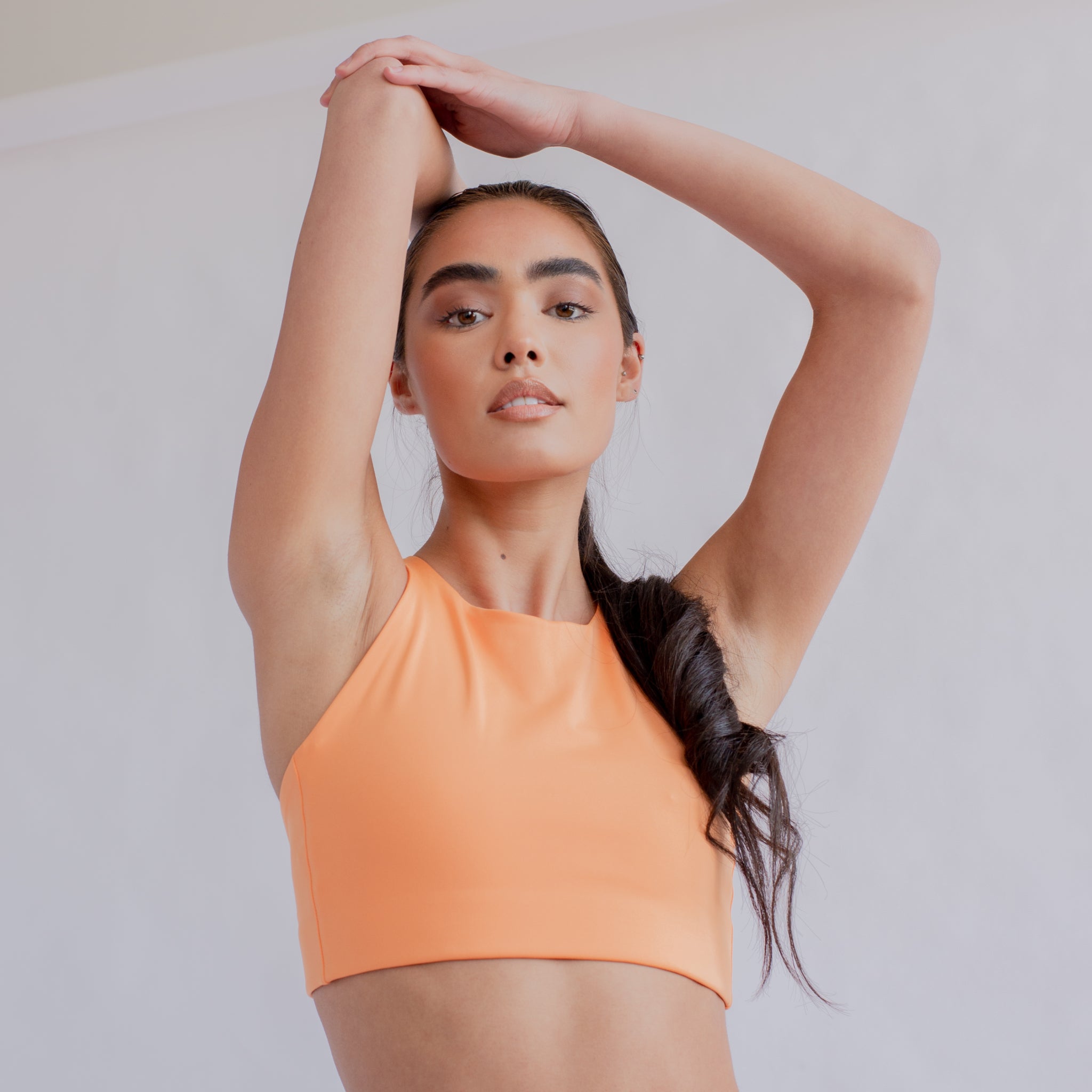 Frau in einem orangefarbenen Oberteil posiert mit erhobenen Armen vor einem hellen Hintergrund.