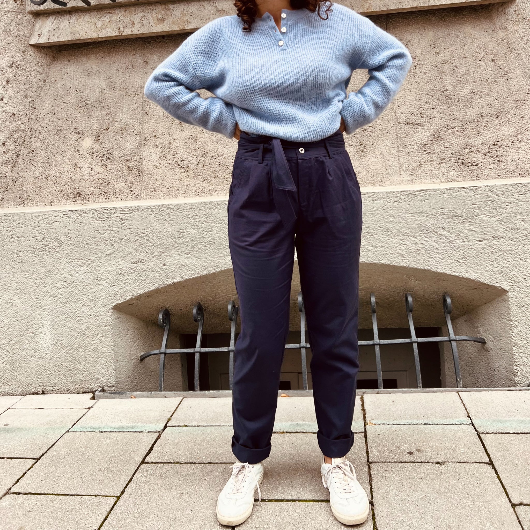 Eine Frau steht vor einer Wand, trägt einen blauen Pullover, dunkle Hose und weiße Sneakers.