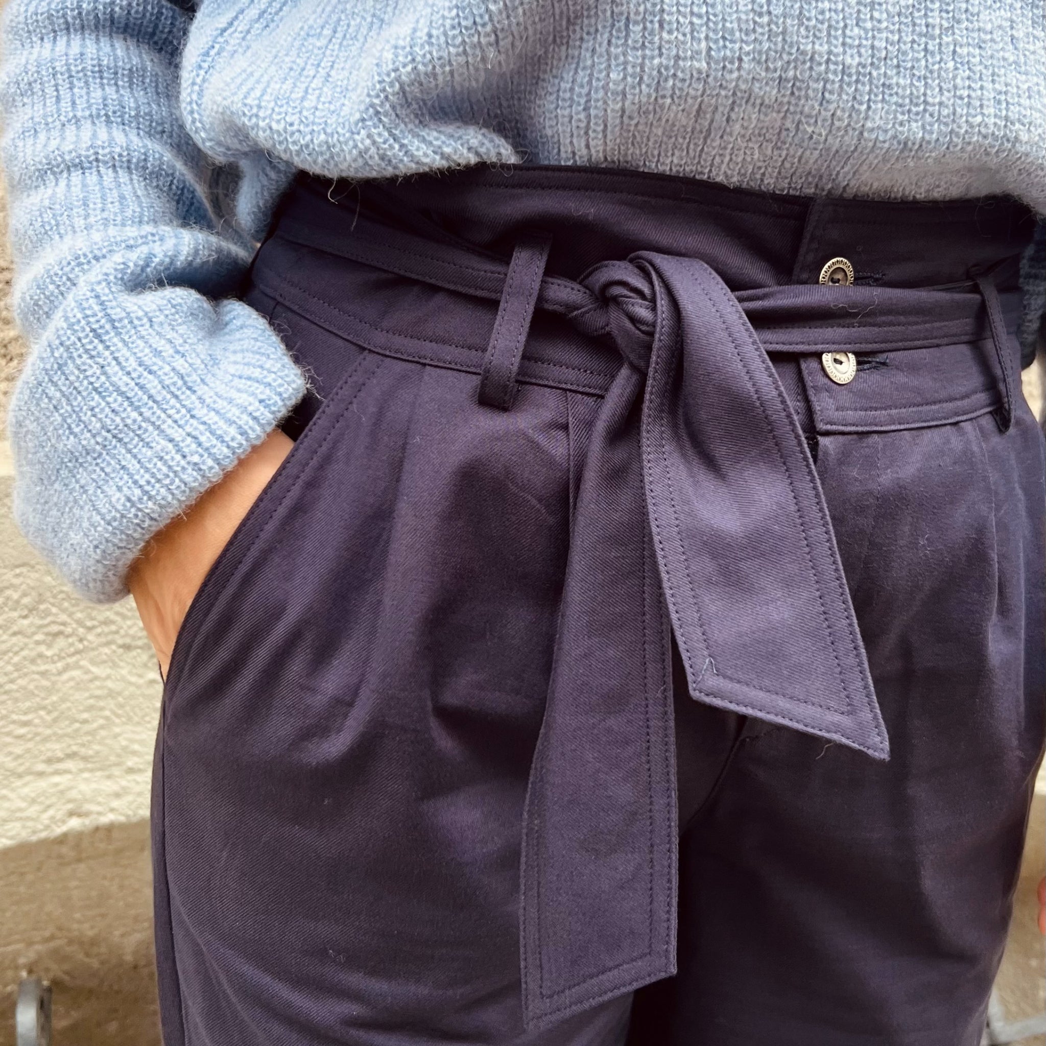 Person trägt eine hellblaue Strickjacke und eine dunkle Hose mit breitem Gürtel, die in der Taille gebunden ist.