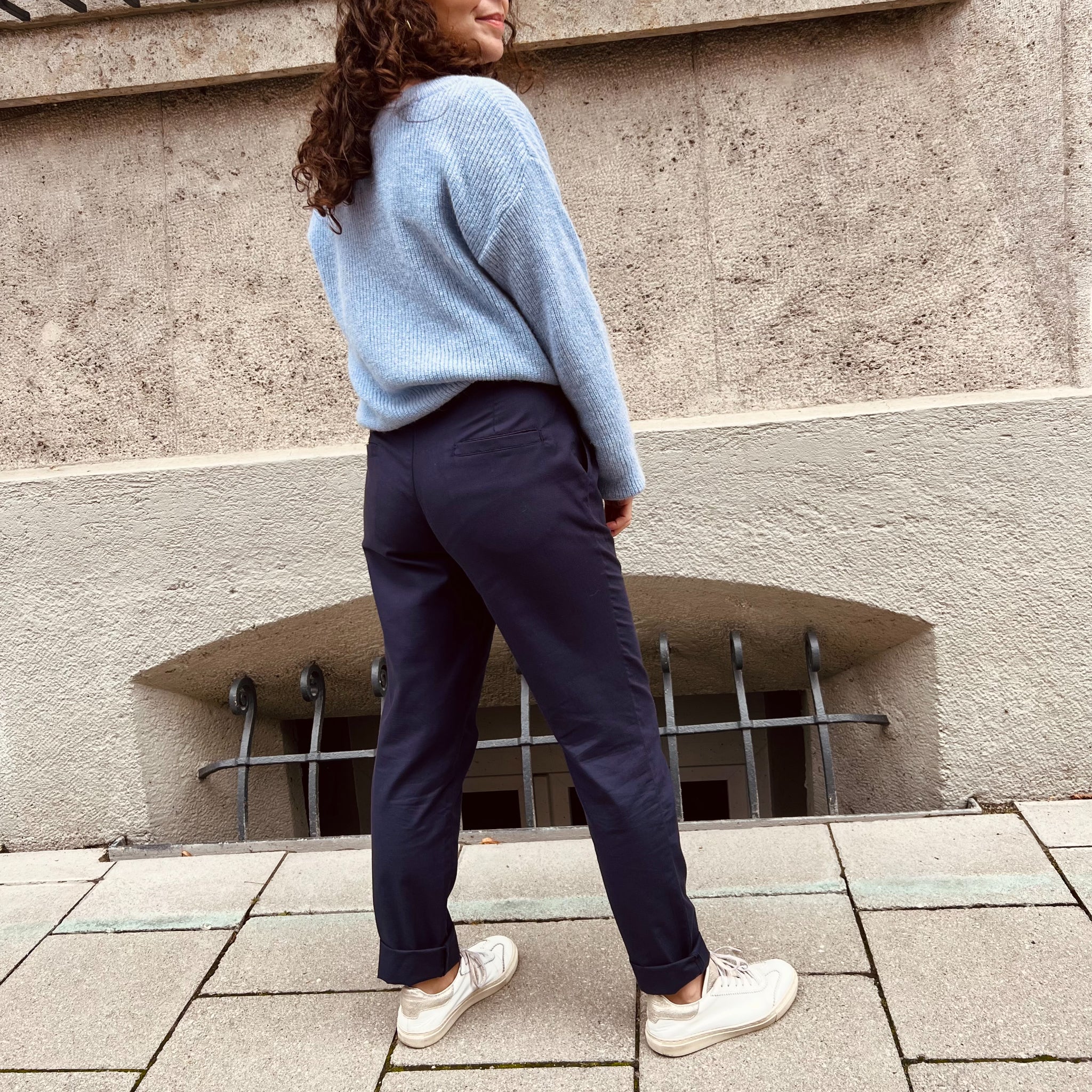 Eine Person steht draußen vor einer Wand und trägt einen blauen Pullover, dunkelblaue Hose und weiße Turnschuhe.