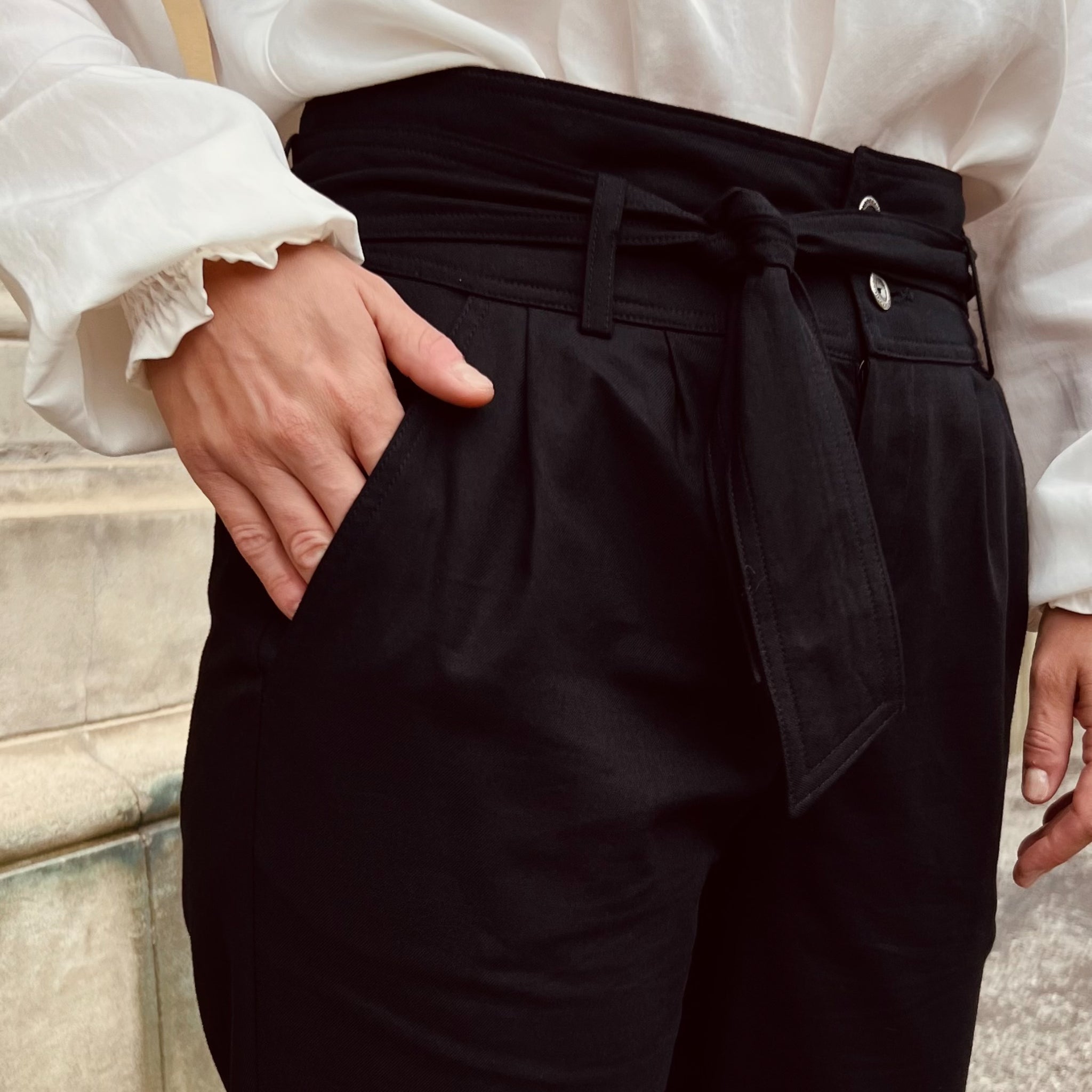 Person trägt schwarze Hose mit Gürtel und weißer Bluse, eine Hand in der Hosentasche.