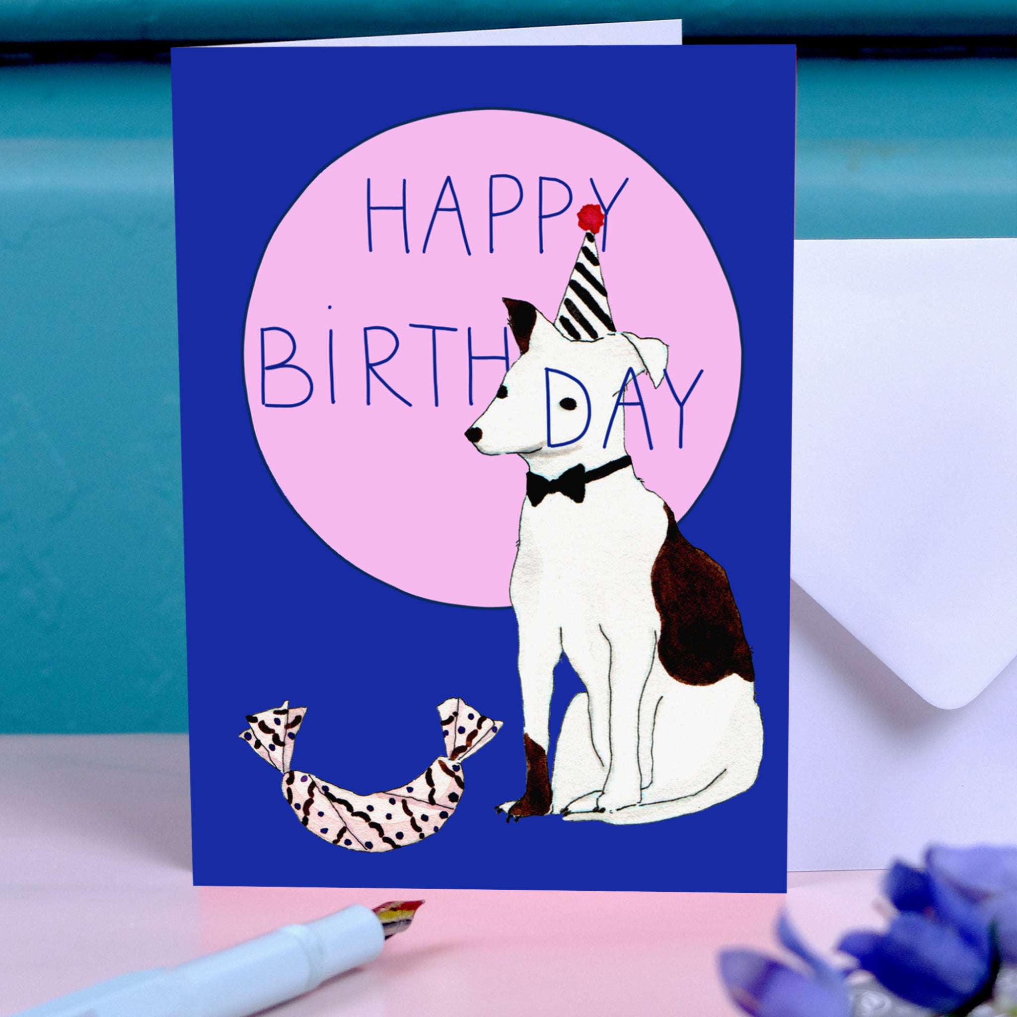 Geburtstagskarte mit Hund in Partyhut und Fliege auf blauem Hintergrund, der "Happy Birthday" in einem rosa Kreis steht.