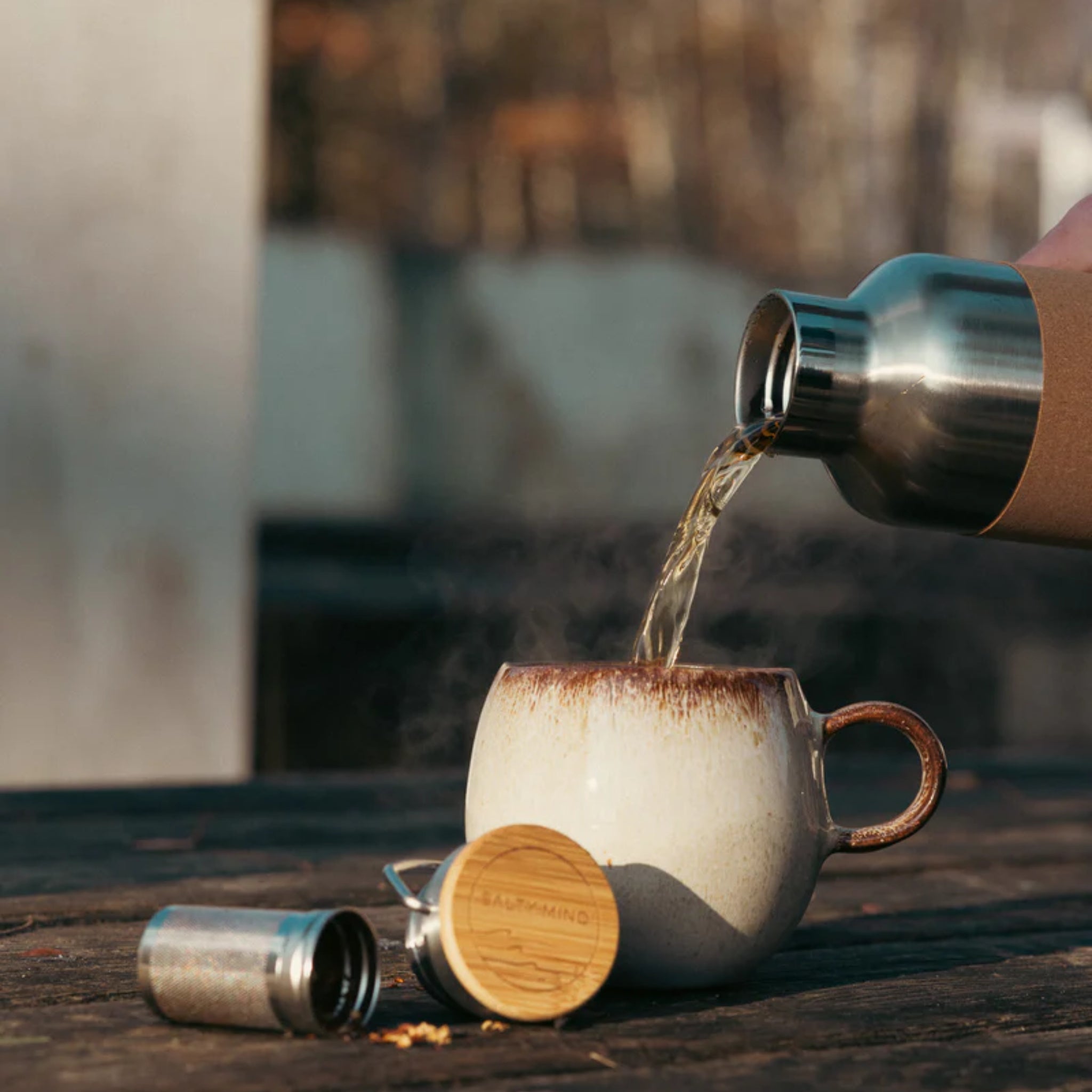 Eine Thermosflasche gießt heißen Tee in eine dampfende Tasse auf einem Holztisch im Freien.