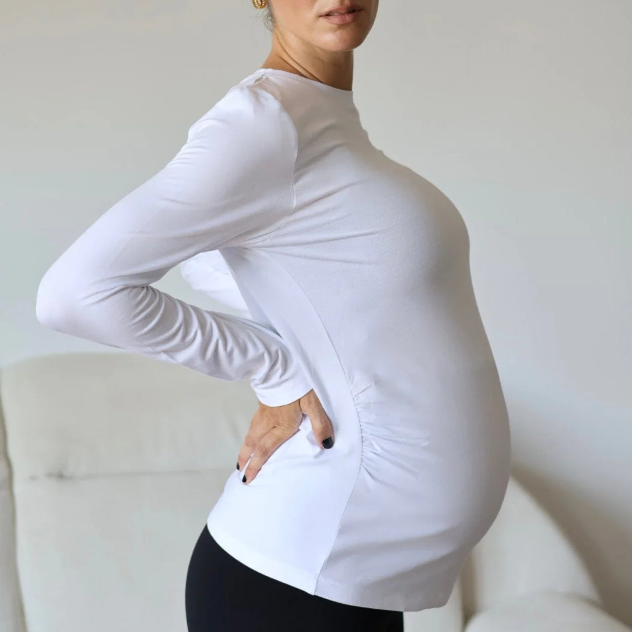 Eine schwangere Frau in einem weißen, langärmeligen Hemd und einer schwarzen Hose steht im Profil, eine Hand auf der Hüfte, um ihren Babybauch zu betonen. Im Hintergrund ist ein helles Sofa zu sehen.