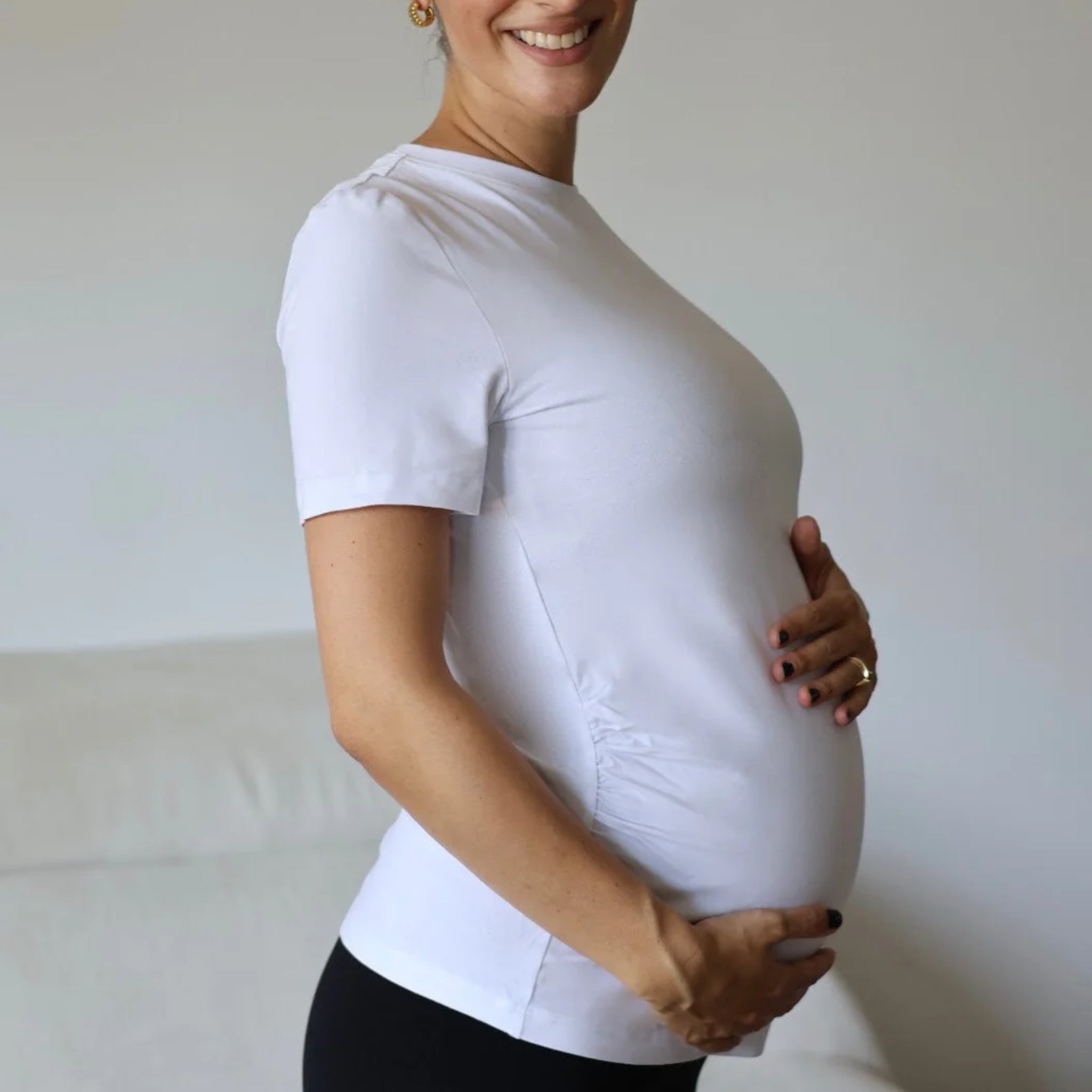 Eine schwangere Frau steht im Profil, lächelt und streichelt mit beiden Händen sanft ihren Babybauch. Sie trägt ein weißes kurzärmeliges Hemd und eine schwarze Hose vor einem neutralen Hintergrund.