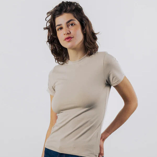 Eine Frau mit kurzen, gewellten braunen Haaren, bekleidet mit einem beigefarbenen T-Shirt und blauen Jeans, steht vor einem schlichten hellgrauen Hintergrund, eine Hand auf die Hüfte gestützt und mit einem neutralen Gesichtsausdruck.