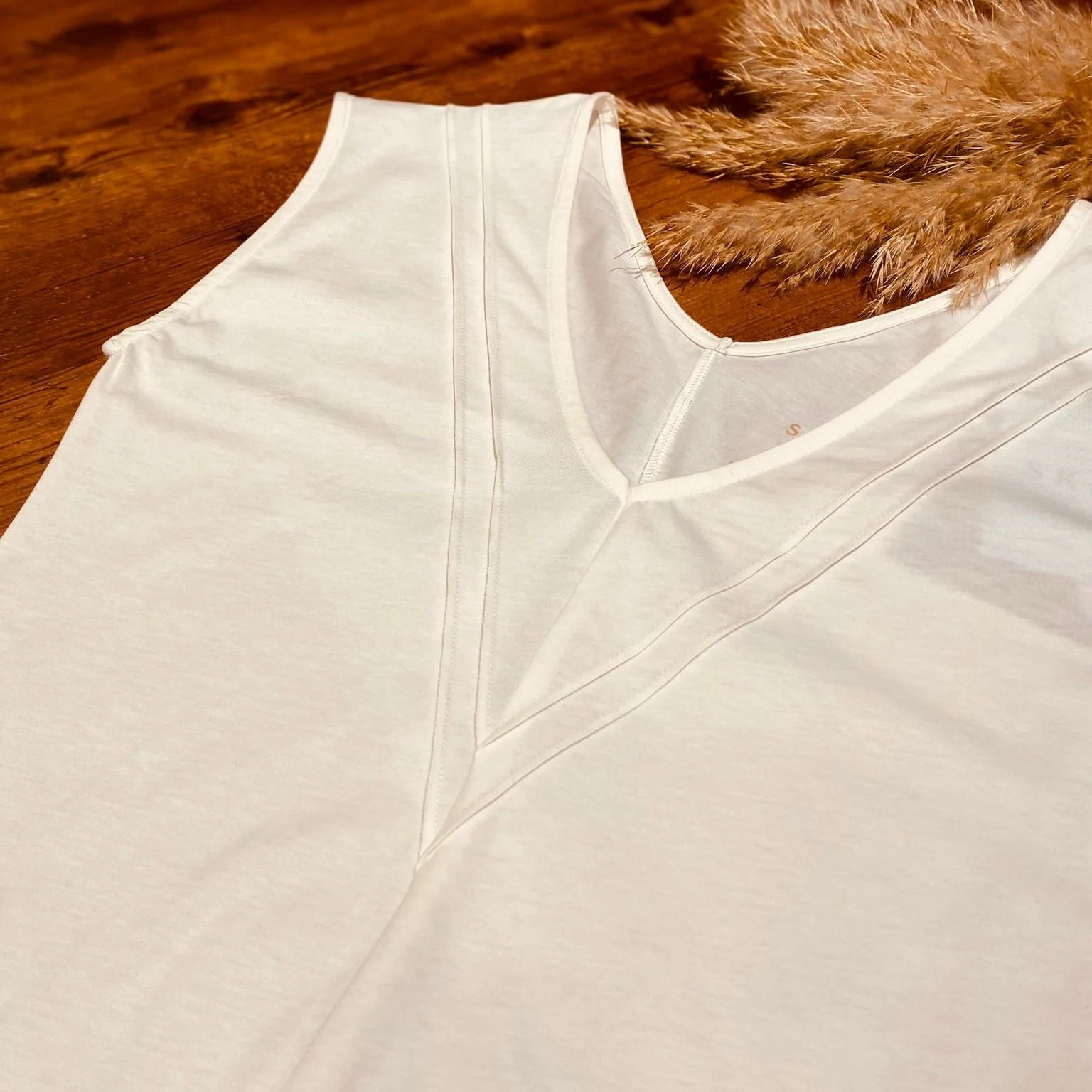 Ein weißes ärmelloses Oberteil mit V-Ausschnitt und Ziernähten liegt flach auf einer hölzernen Unterlage, auf der in der Nähe des Ausschnitts getrocknetes Pampasgras liegt.