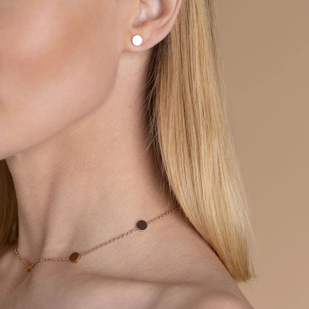 Nahaufnahme einer Frau mit glattem blondem Haar, die einen kleinen runden goldenen Ohrstecker und eine zarte goldene Halskette mit passenden runden Anhängern vor einem beigen Hintergrund trägt.