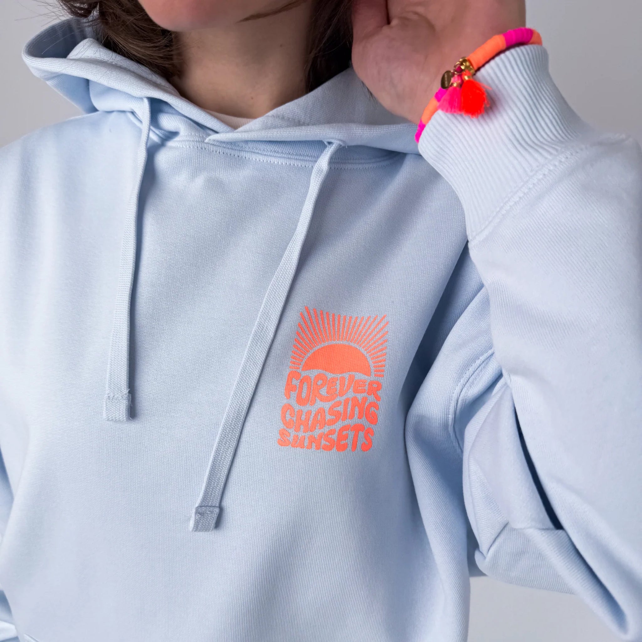 Eine Person, die einen hellblauen Kapuzenpullover mit einer pink-orangenen Sonnengrafik und der Aufschrift FOREVER CHASING SUNSETS sowie ein neonpinkes Armband mit Quasten am Handgelenk trägt.