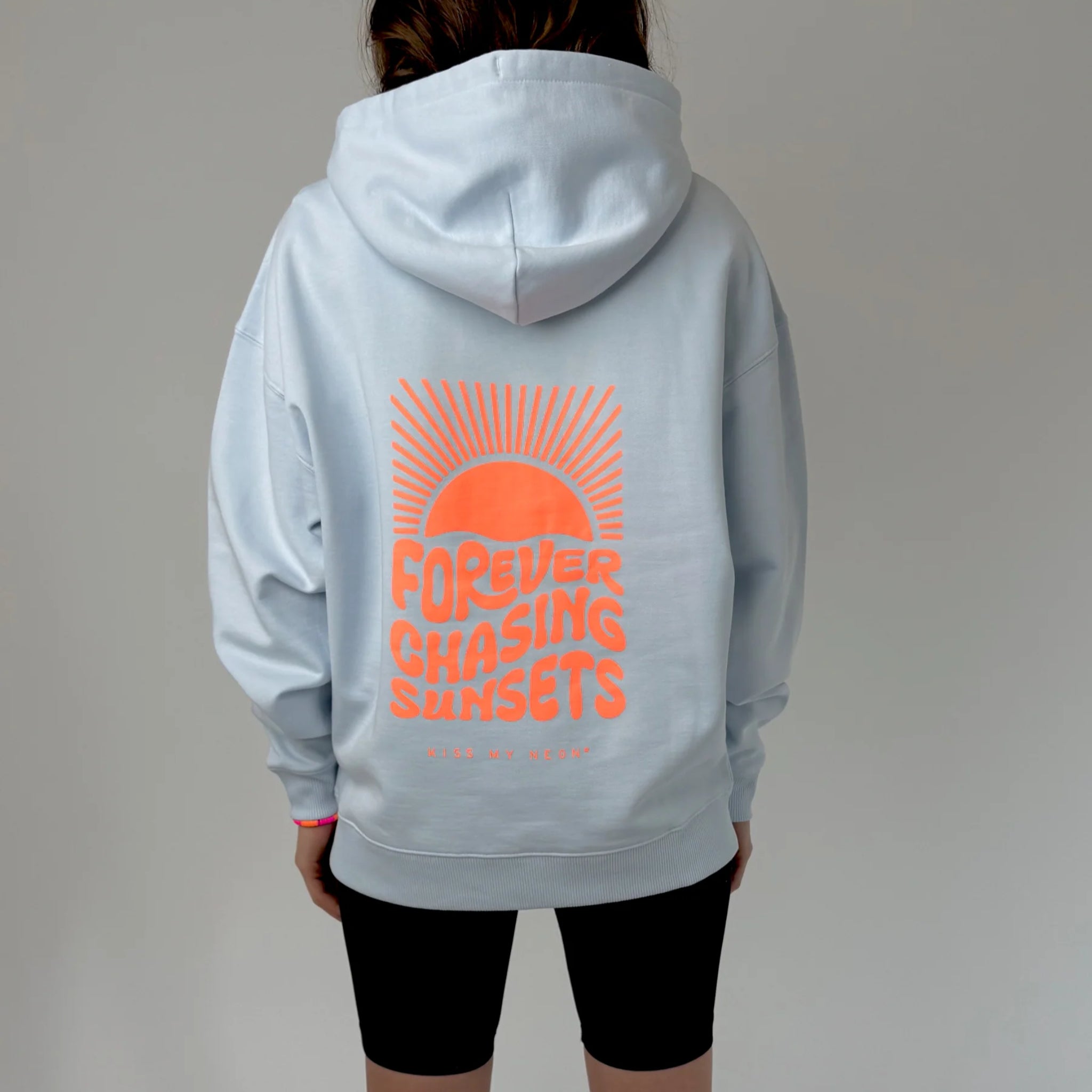 Die Person trägt einen hellblauen Kapuzenpullover mit einer Retro-Grafik einer orangefarbenen Sonne und Strahlen auf dem Rücken, mit der Aufschrift Forever Chasing Sunsets und kleinerem Text darunter. Die Person trägt außerdem schwarze Shorts.