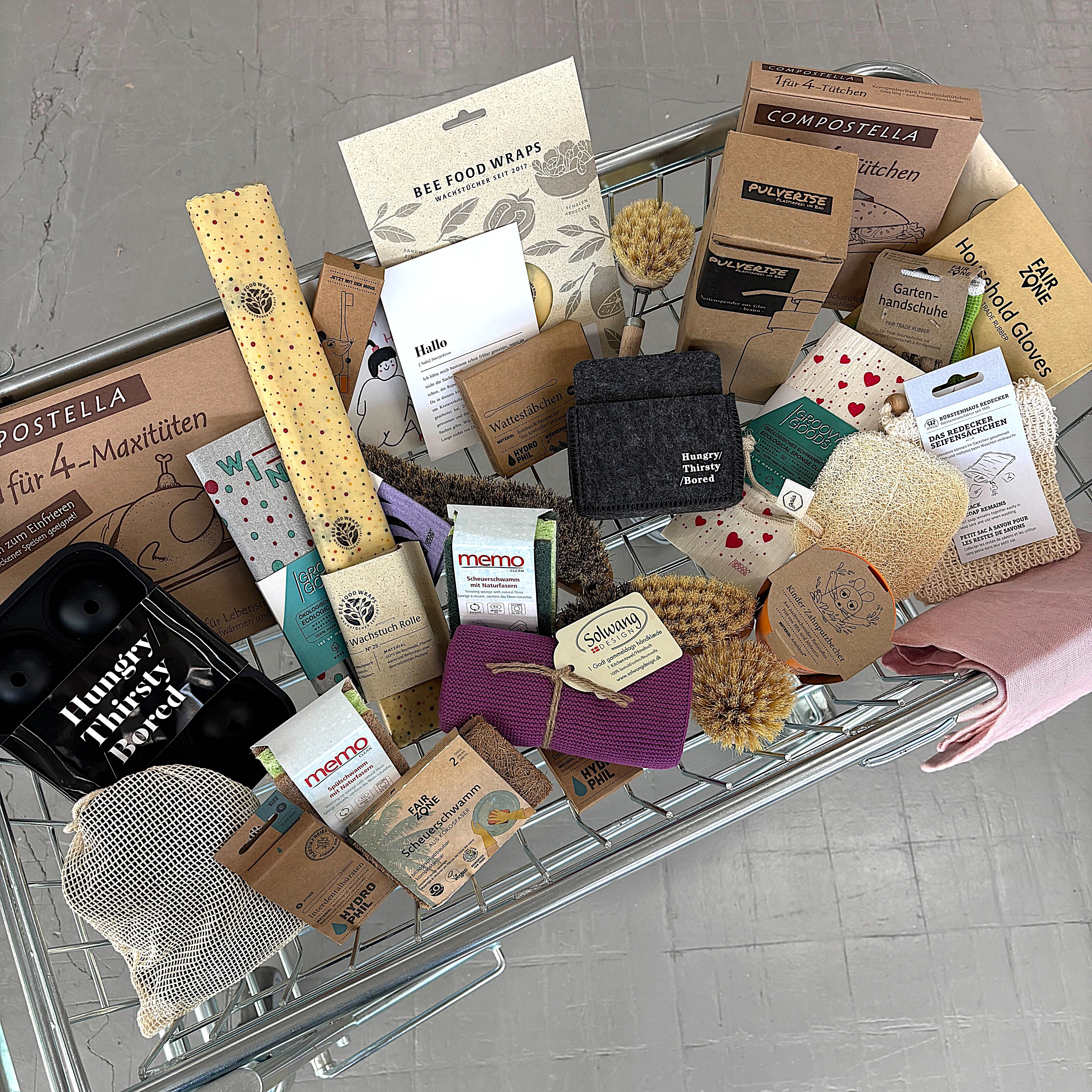 Ein Einkaufswagen, gefüllt mit verschiedenen umweltfreundlichen und nachhaltigen Haushaltsprodukten, darunter Bienenwachswickel, Schwämme, Bürsten, kompostierbare Taschen, wiederverwendbare Tücher und Naturseifen, ordentlich auf einem grauen Boden angeordnet.