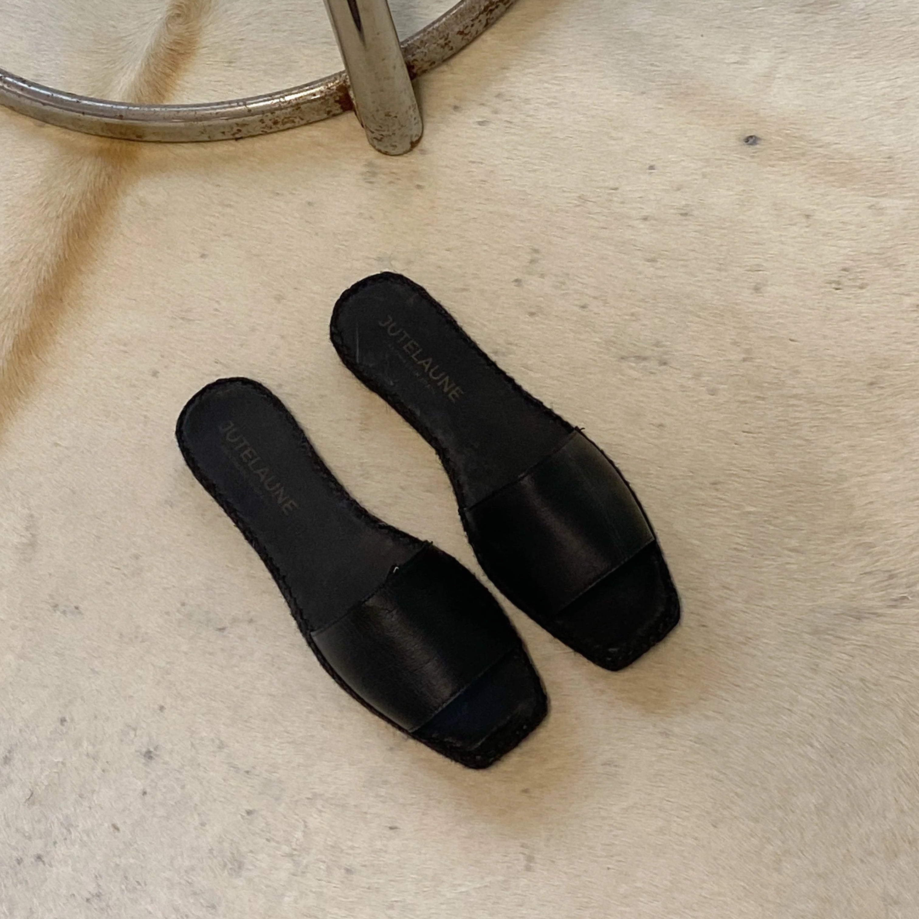 Ein Paar schwarzer, offener Pantoletten liegt auf einem hellen, strukturierten Teppich neben einem modernen runden Beistelltisch aus Glas und Chrom.