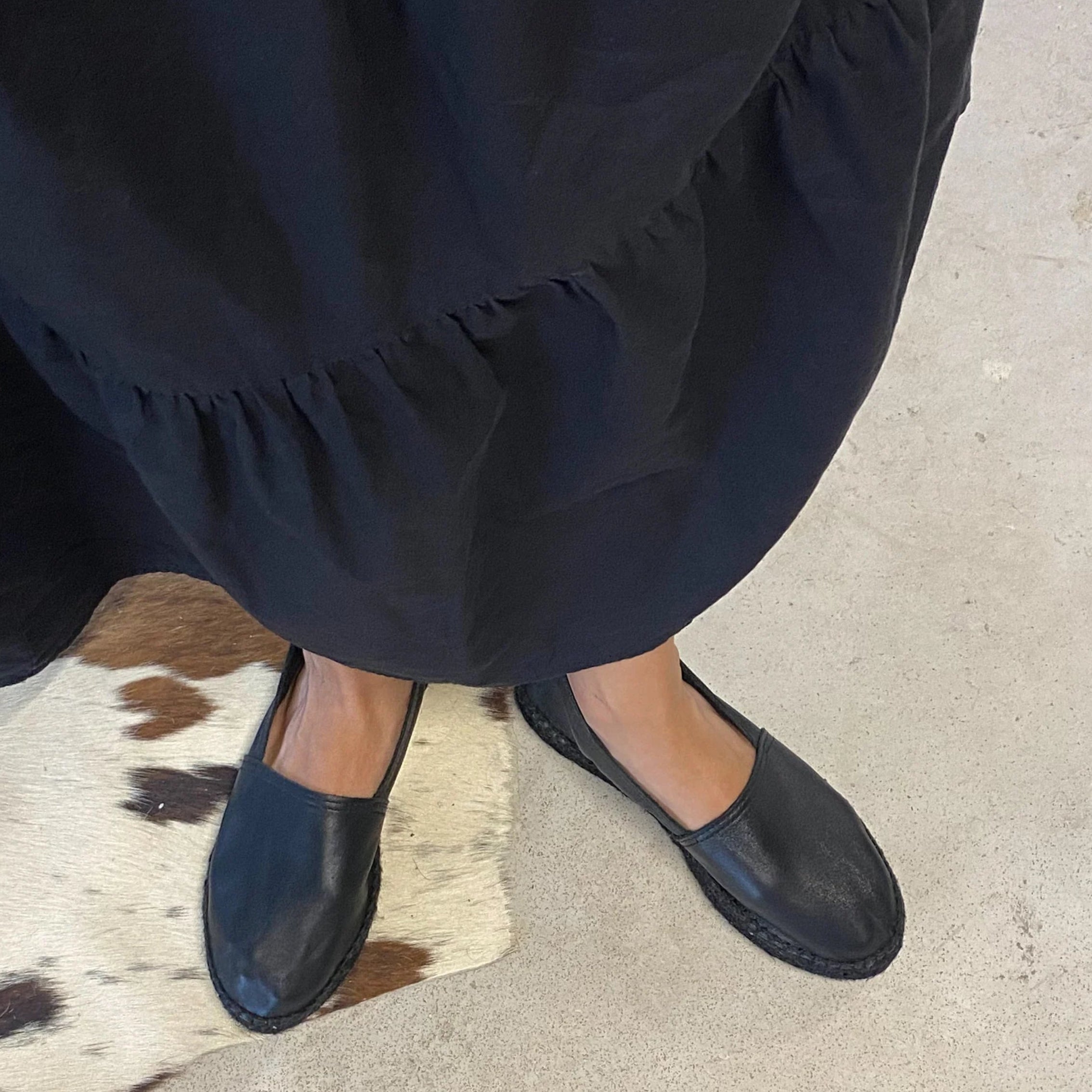 Eine Person in einem langen, schwarzen, gerüschten Kleid und schwarzen Slippern steht auf einem hellen Boden neben einem gefleckten Kuhfellteppich.