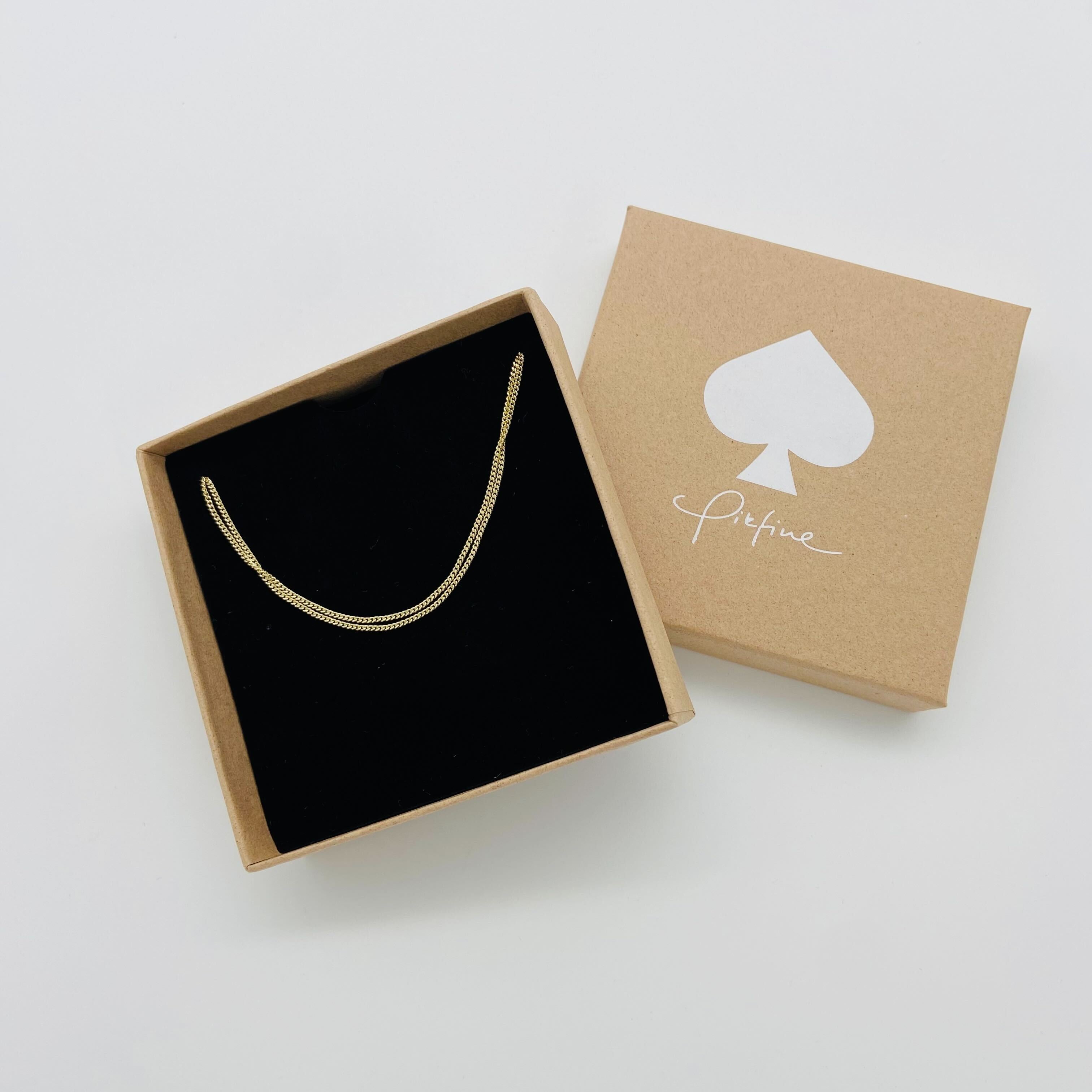 Goldene Halskette in einer schwarzen Schachtel mit einem braunen Deckel, verziert mit einem weißen Pik-Symbol.