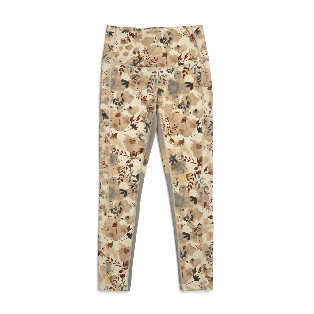 Eine Person hält ein Paar beigefarbene Leggings mit einem Blumenmuster vor einer weißen Wand hoch. Der Kopf der Person befindet sich außerhalb des Bildausschnitts, und sie trägt eine dunkle Jacke und eine grüne Hose.