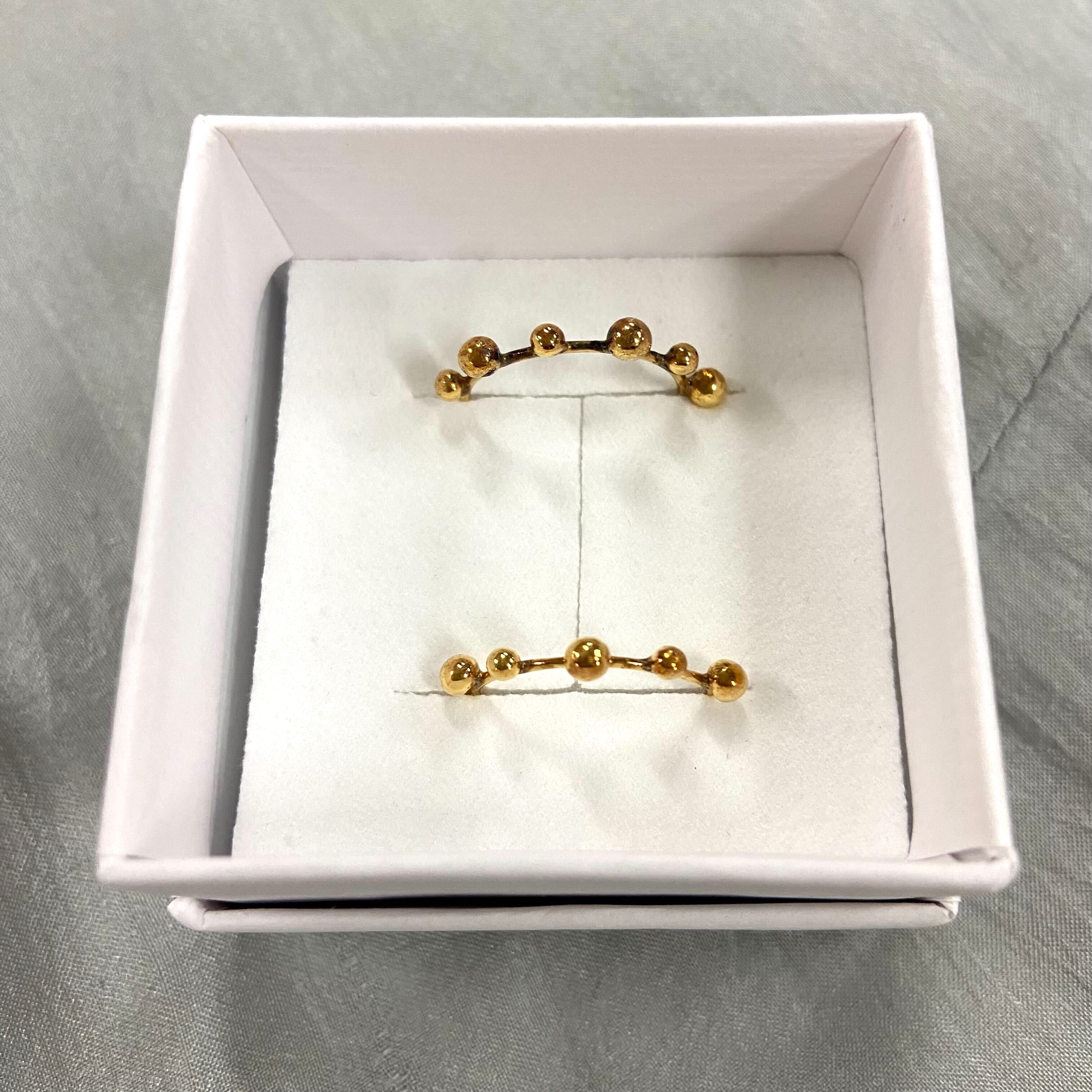 Zwei goldene, durch Perlen verbundene Ohrstecker liegen in einer weißen Geschenkbox auf einem hellgrauen Hintergrund.