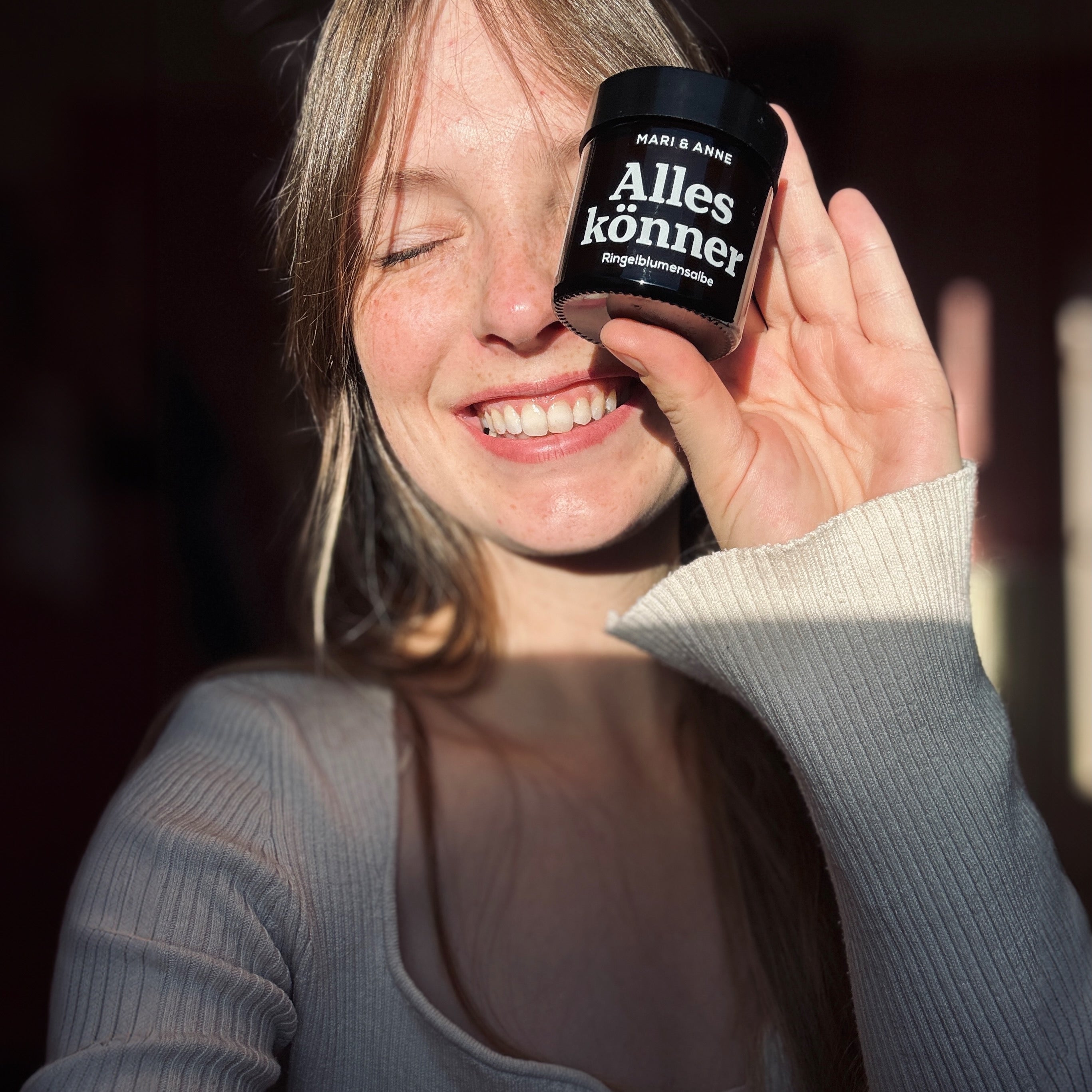 Eine lächelnde Frau hält ein Glas mit der Aufschrift "Alleskönner Ringelblumensalbe" in der Hand.