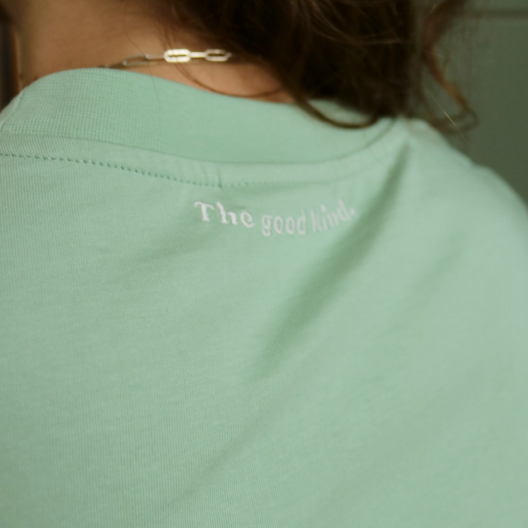 Rückansicht eines grünen Shirts mit dem weißen Schriftzug "The good kind." am Nacken, getragen von einer Person mit Halskette.