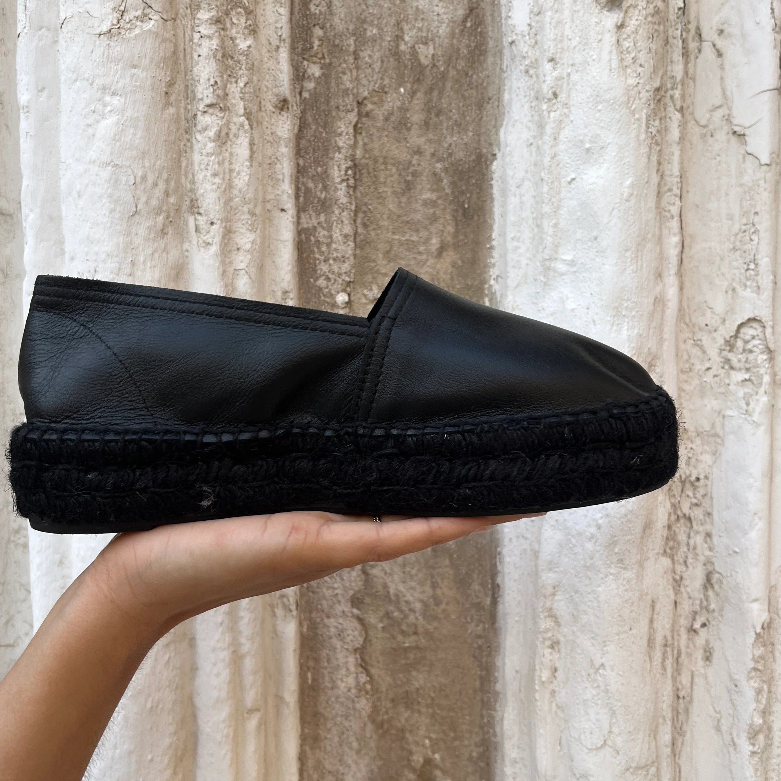 Eine Hand hält einen schwarzen Espadrille-Schuh aus Leder mit einer dicken schwarzen gewebten Sohle vor einer strukturierten weißen und braunen Steinmauer hoch.