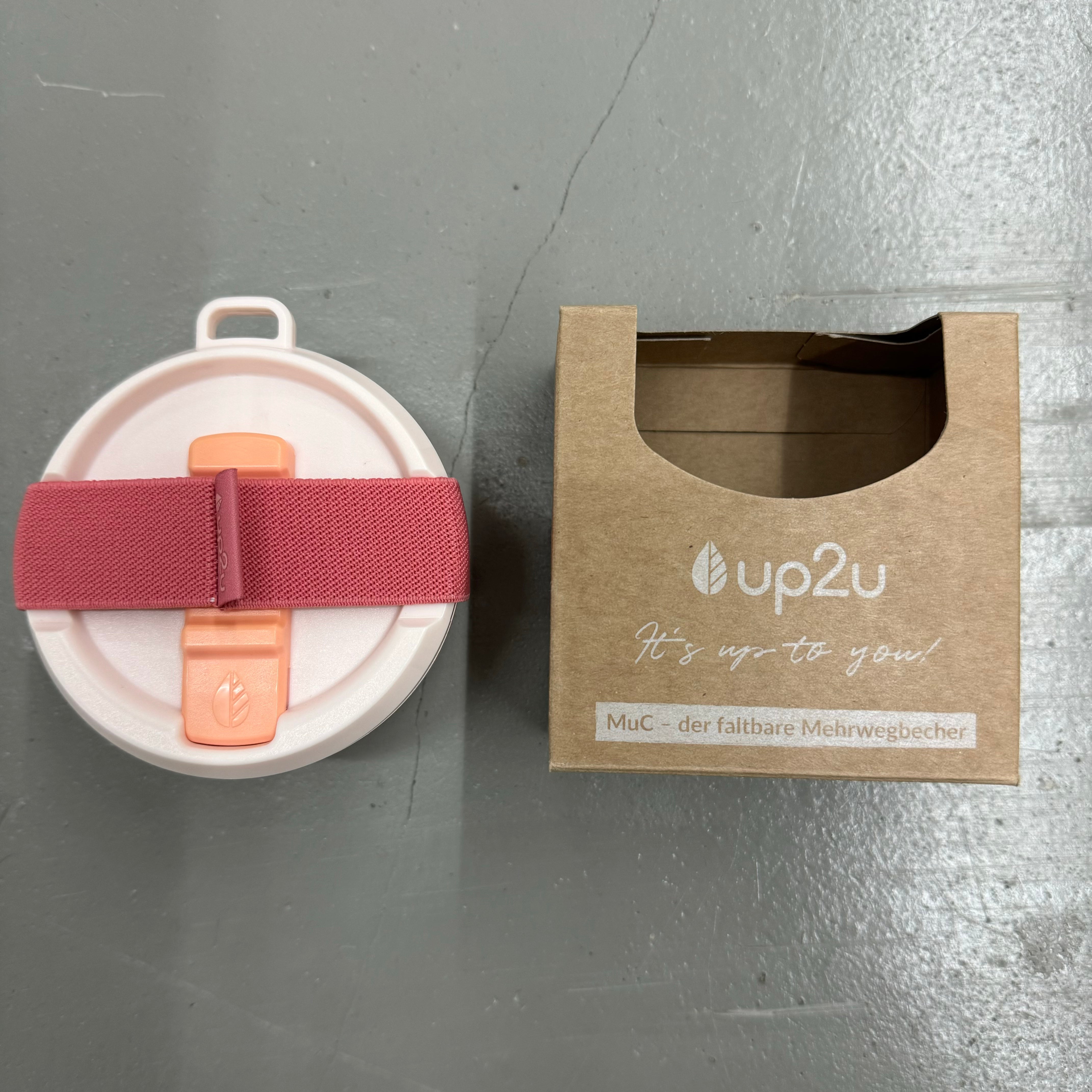Ein zusammenklappbarer Mehrwegbecher mit pinkem Band neben einer braunen Verpackung mit der Aufschrift "up2u".