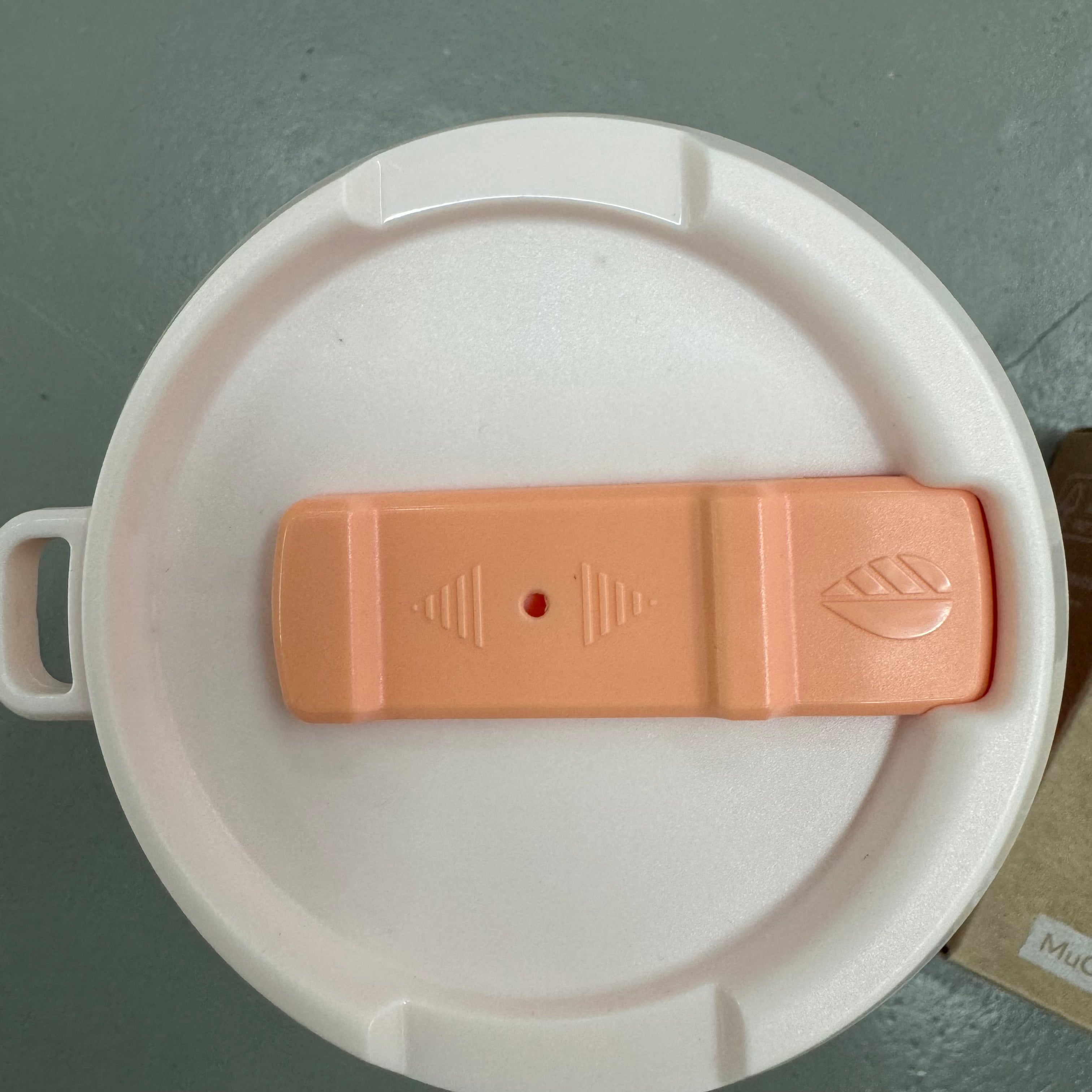 Runder weißer Deckel mit orangefarbenem Schieberegler und Griff auf grauer Oberfläche.