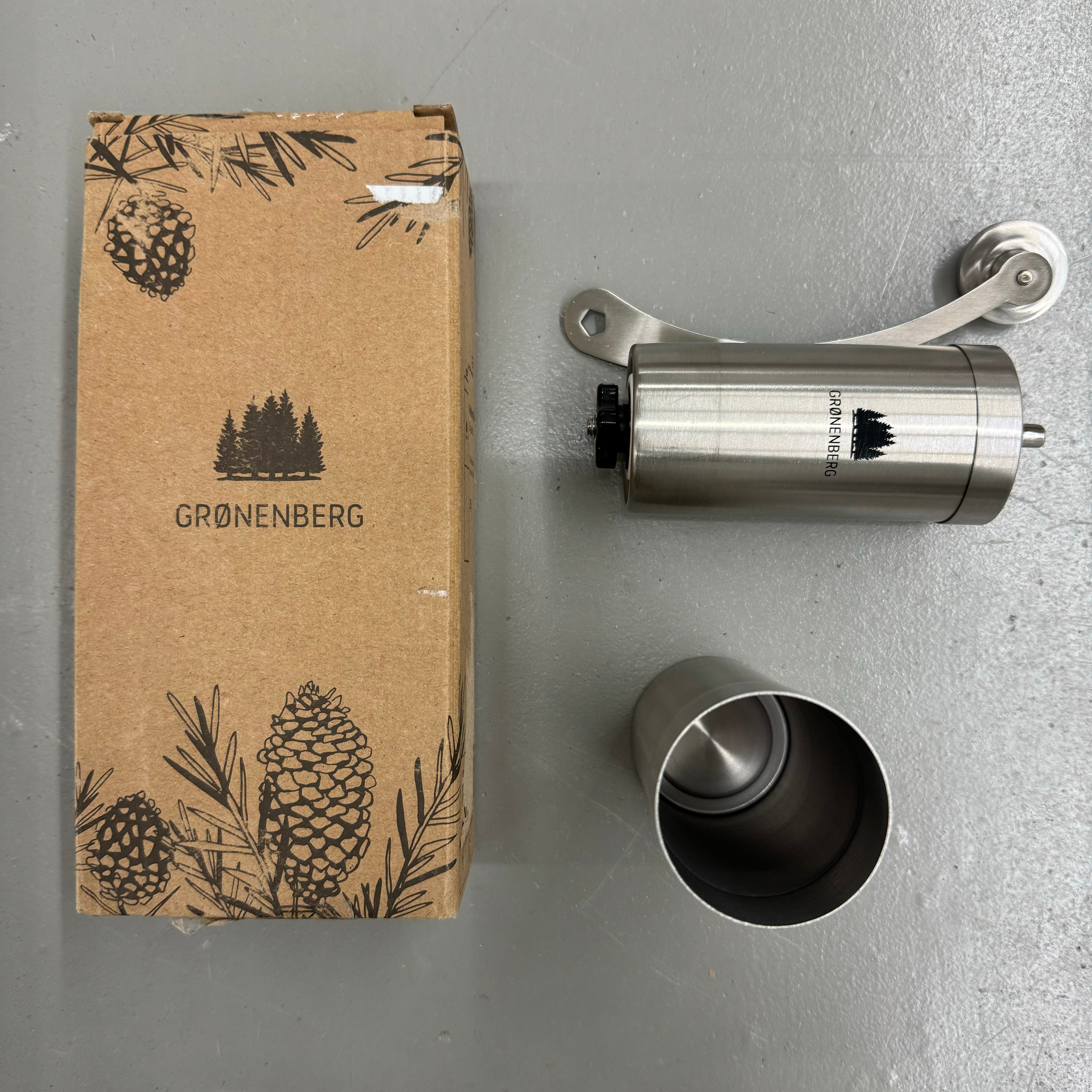 Ein Karton und eine Edelstahl-Kaffeemühle mit dem Logo "GRØNENBERG" und einem Kiefernbaum-Design auf grauem Hintergrund.